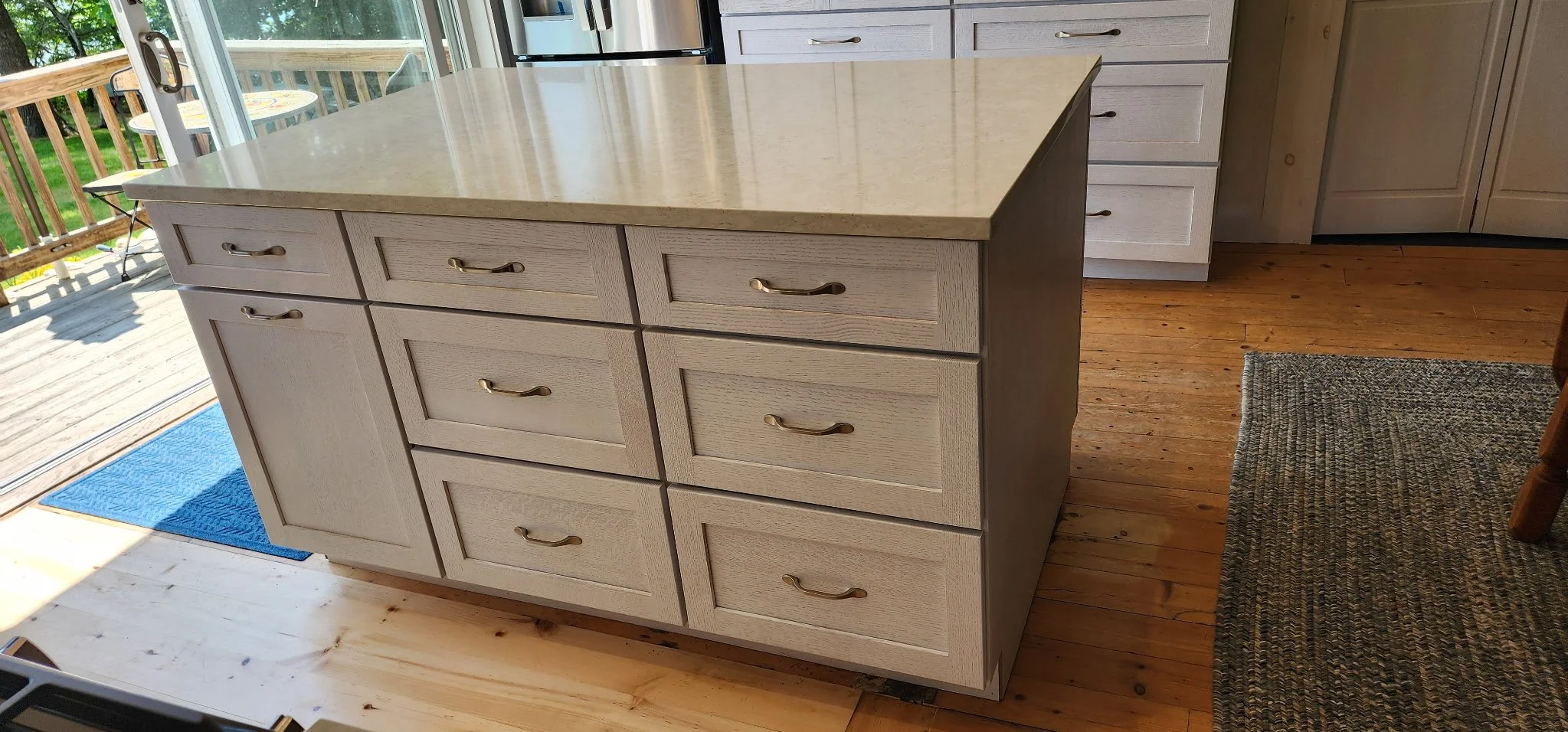 A kitchen island with multiple drawers and a granite countertop, situated on a wooden floor near a sliding glass door leading to an outdoor deck.