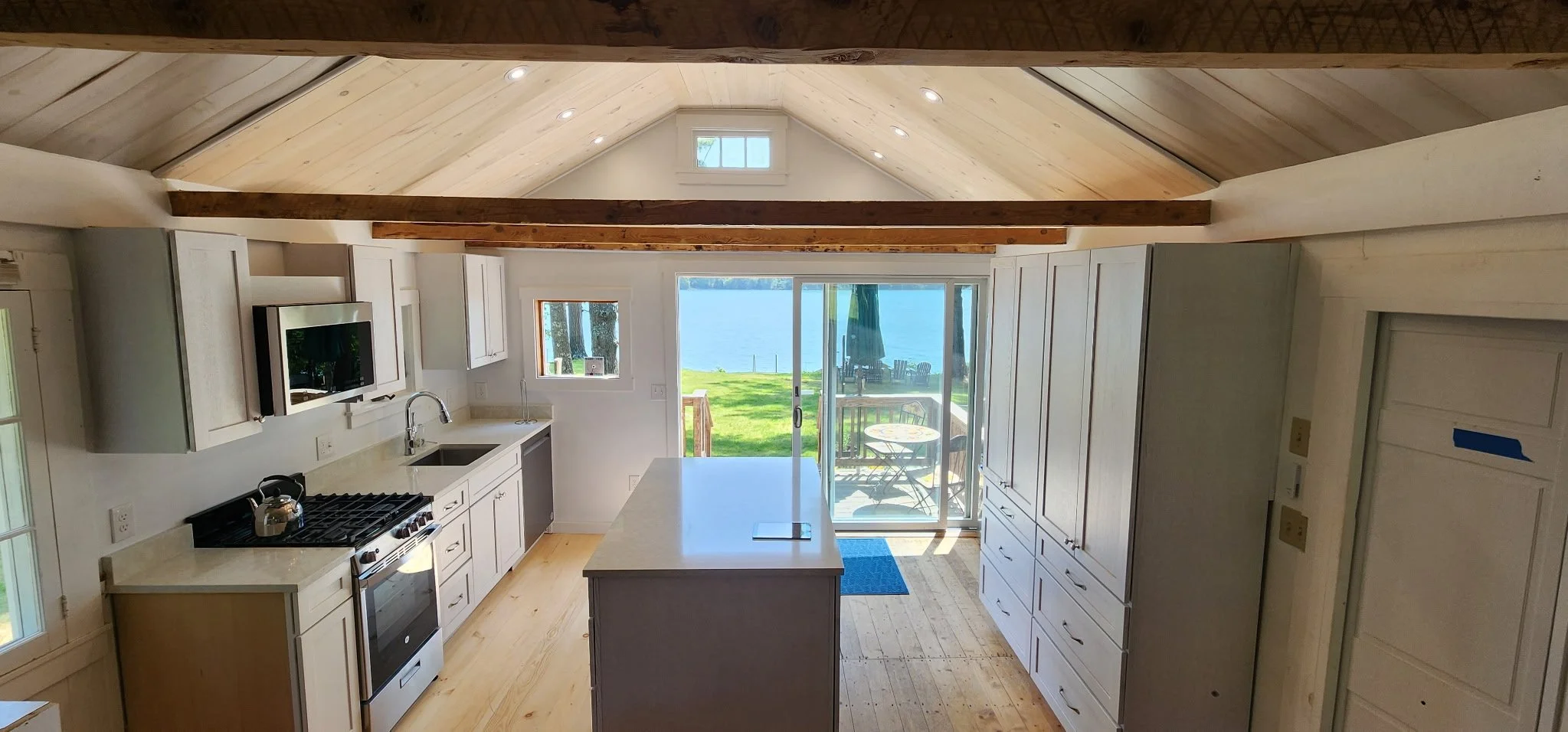 A kitchen with white cabinets, a white countertop, and a central island. There is a stove with a kettle on it, a sink, and a small television mounted on the wall. Large glass sliding doors open to a patio with outdoor seating, overlooking a grassy ya