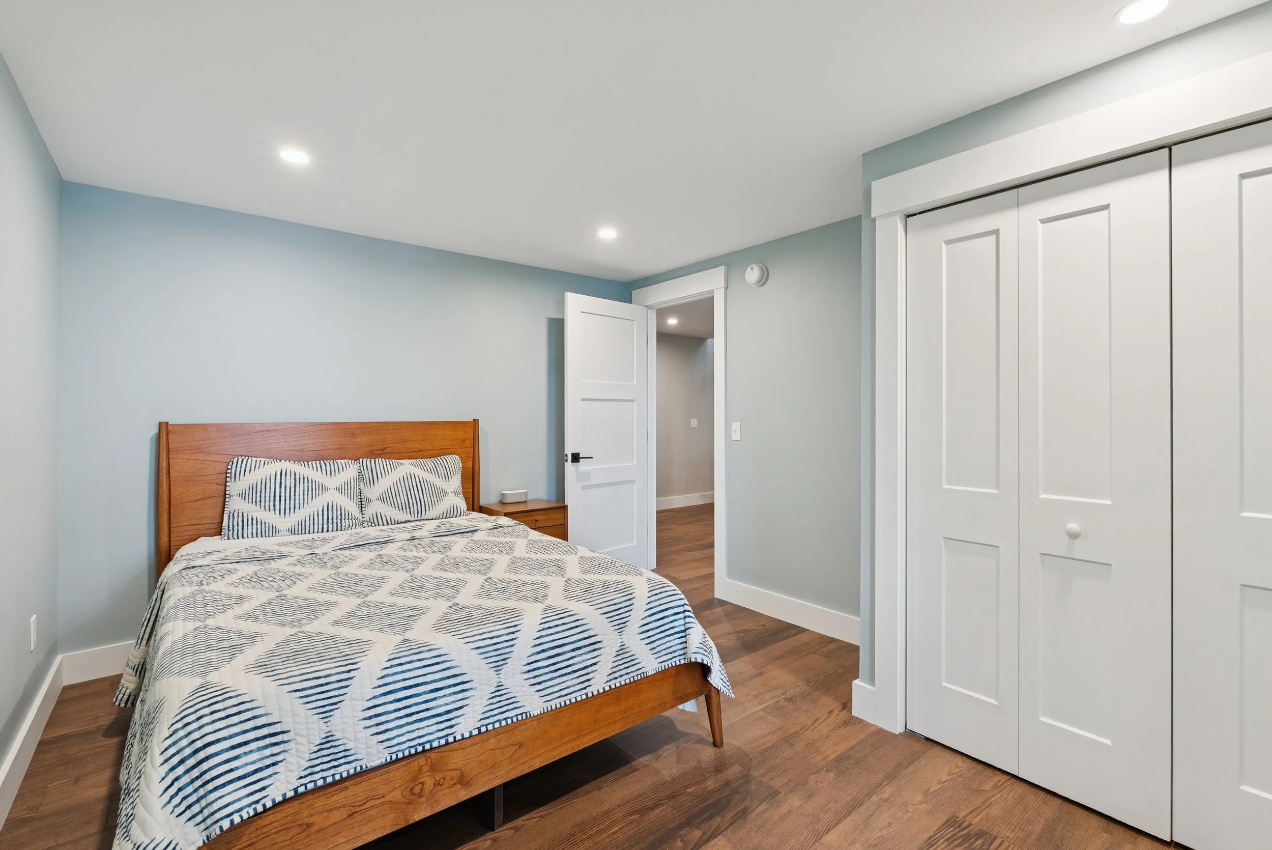A bedroom with light blue walls, a bed with a wooden headboard, a patterned bedspread, and matching pillow, a nightstand, and a white closet door. There are ceiling lights and an open door leading to another room.