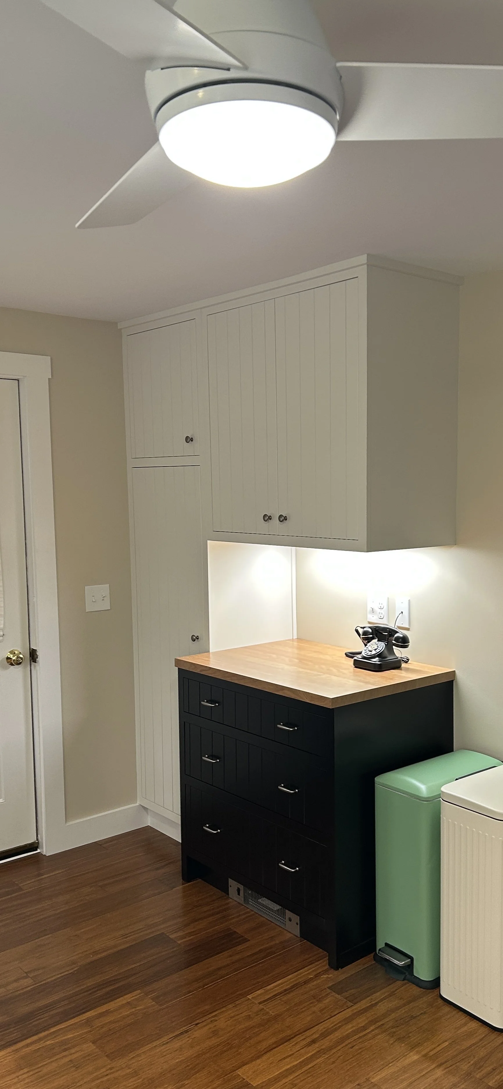 A kitchen corner with a ceiling fan, white cabinets, a wooden countertop, a black spice drawer, a vintage rotary phone, a mint green trash can, and a white cabinet or appliance.