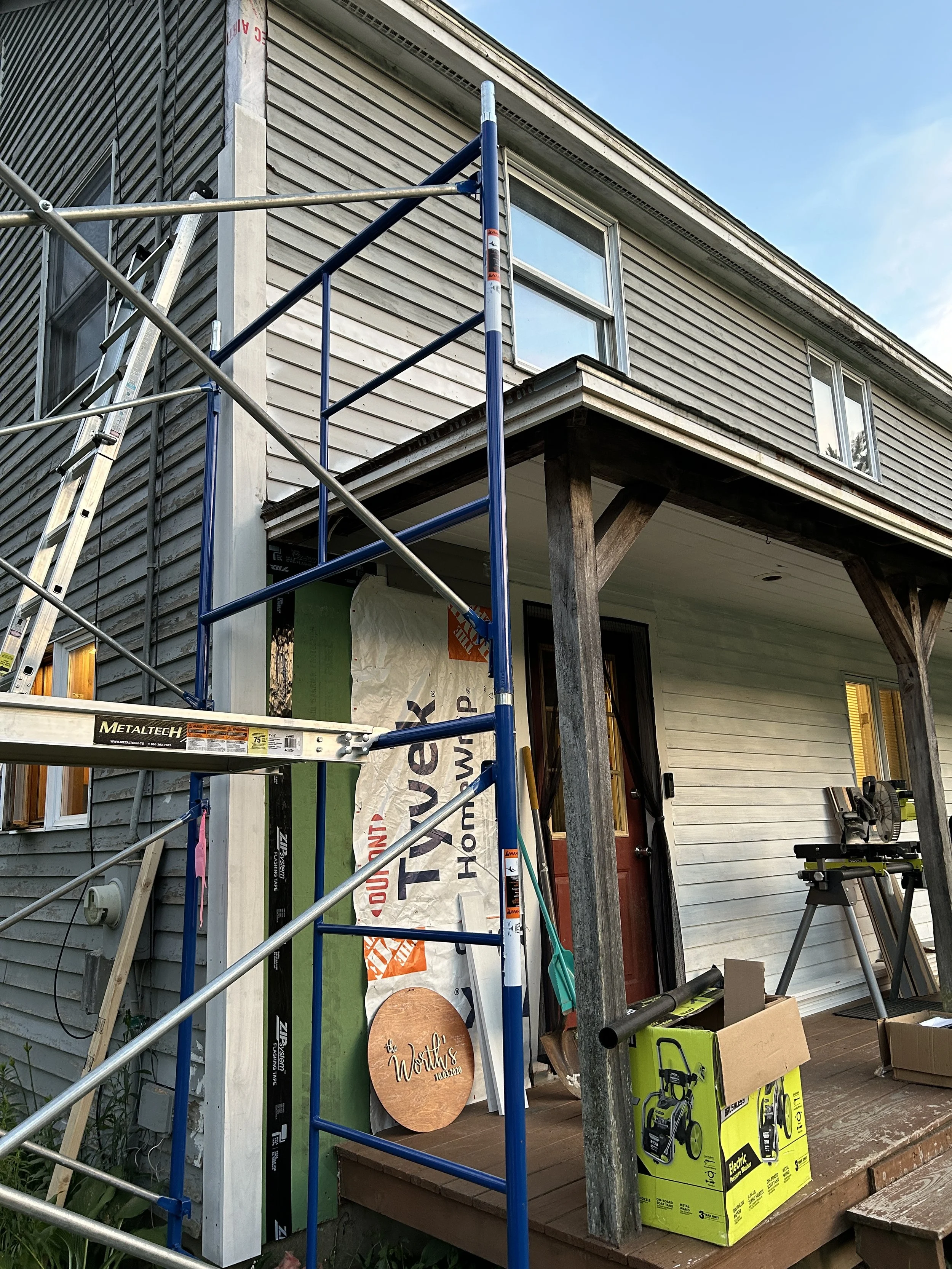 A house exterior under renovation with scaffolding, showing a second story with horizontal siding, windows, and construction materials, including a yellow box and green toolbox on the porch.