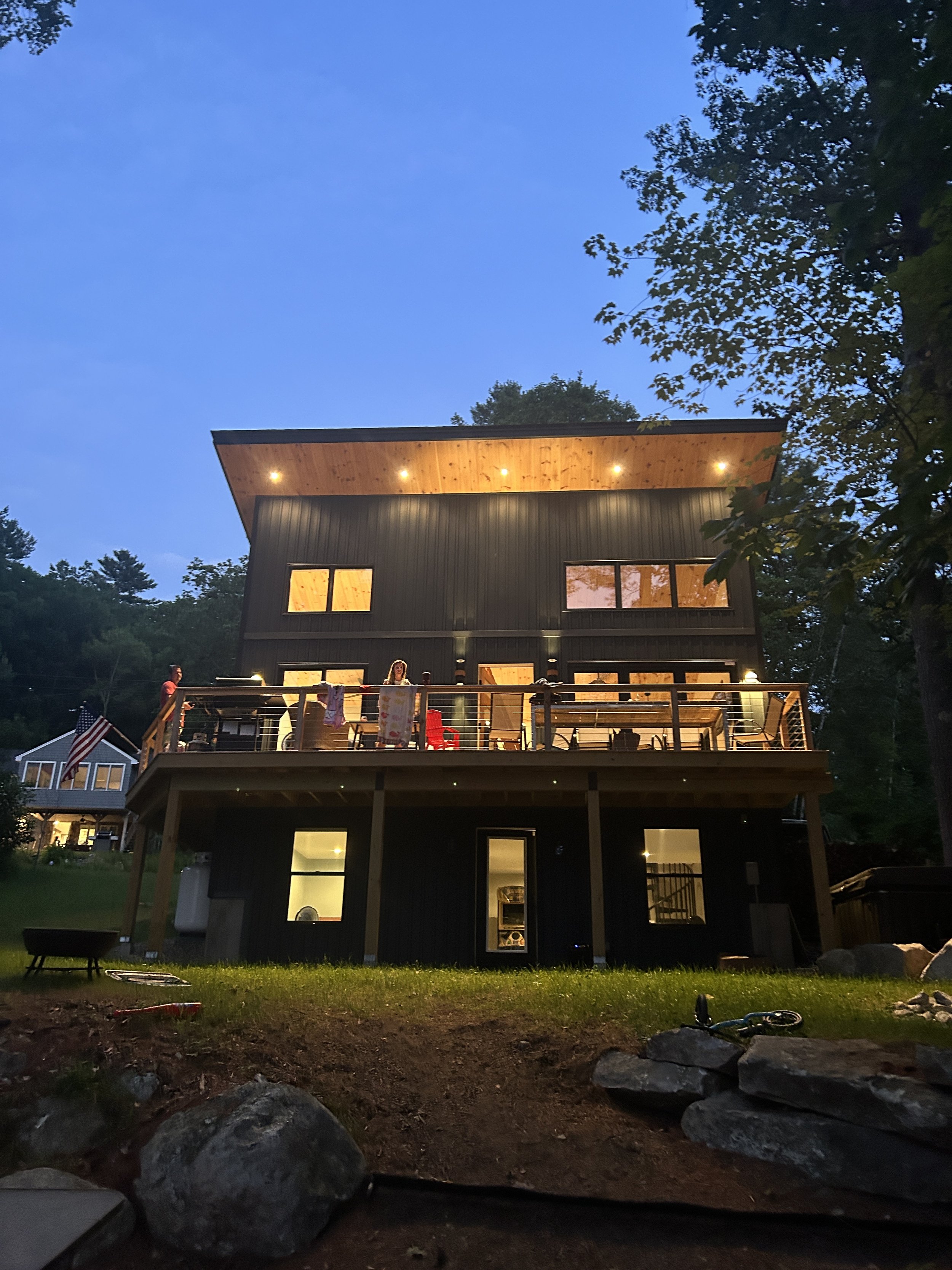 A two-story modern house at dusk with warm lighting inside. The house has black vertical siding, large windows on both floors, and a wooden roof overhang with recessed lights. There is a balcony on the second floor with a person standing near the rai
