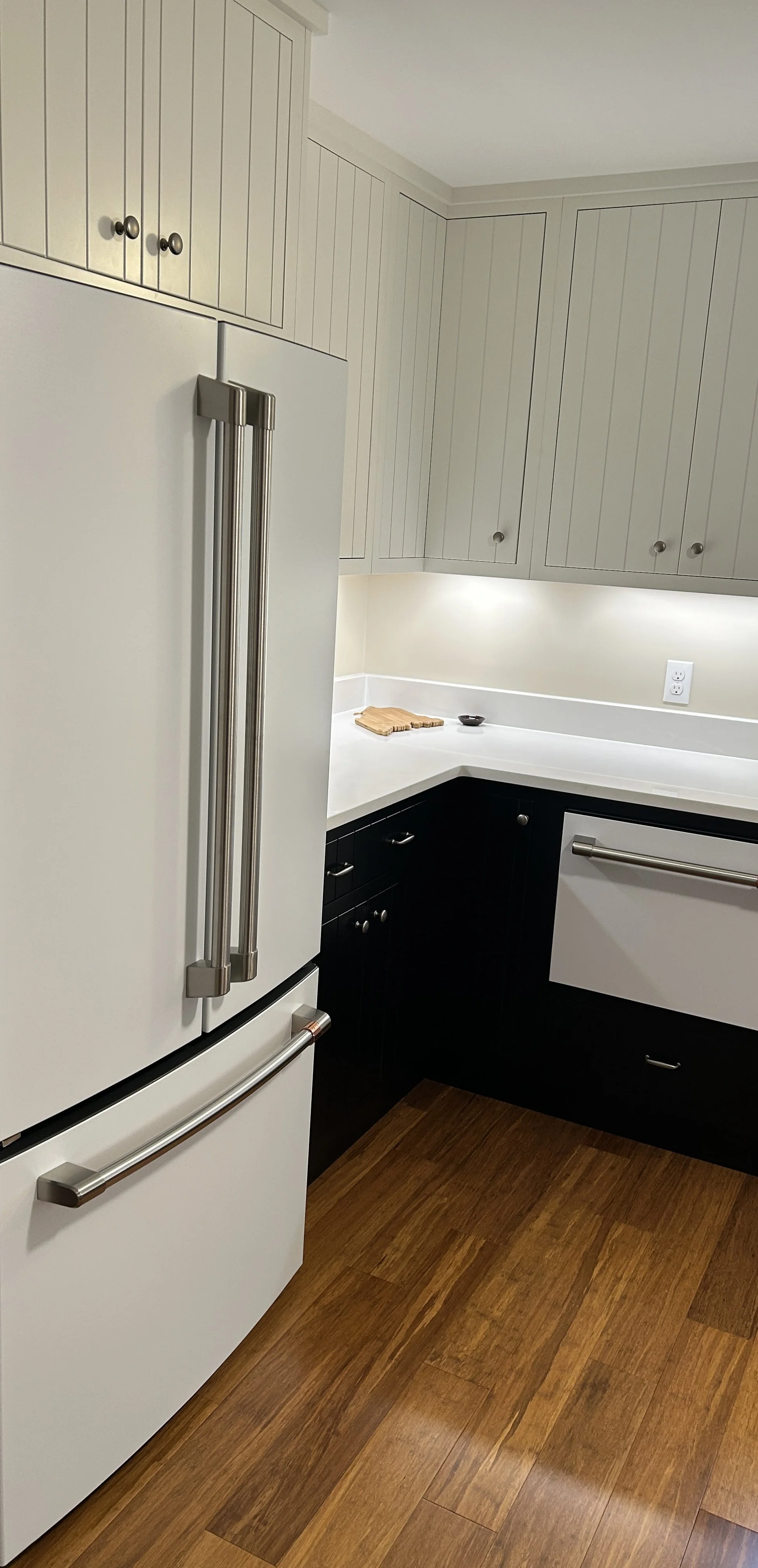 Kitchen with white refrigerator, white and dark cabinets, wooden floor, and white countertop illuminated by under cabinet lighting.