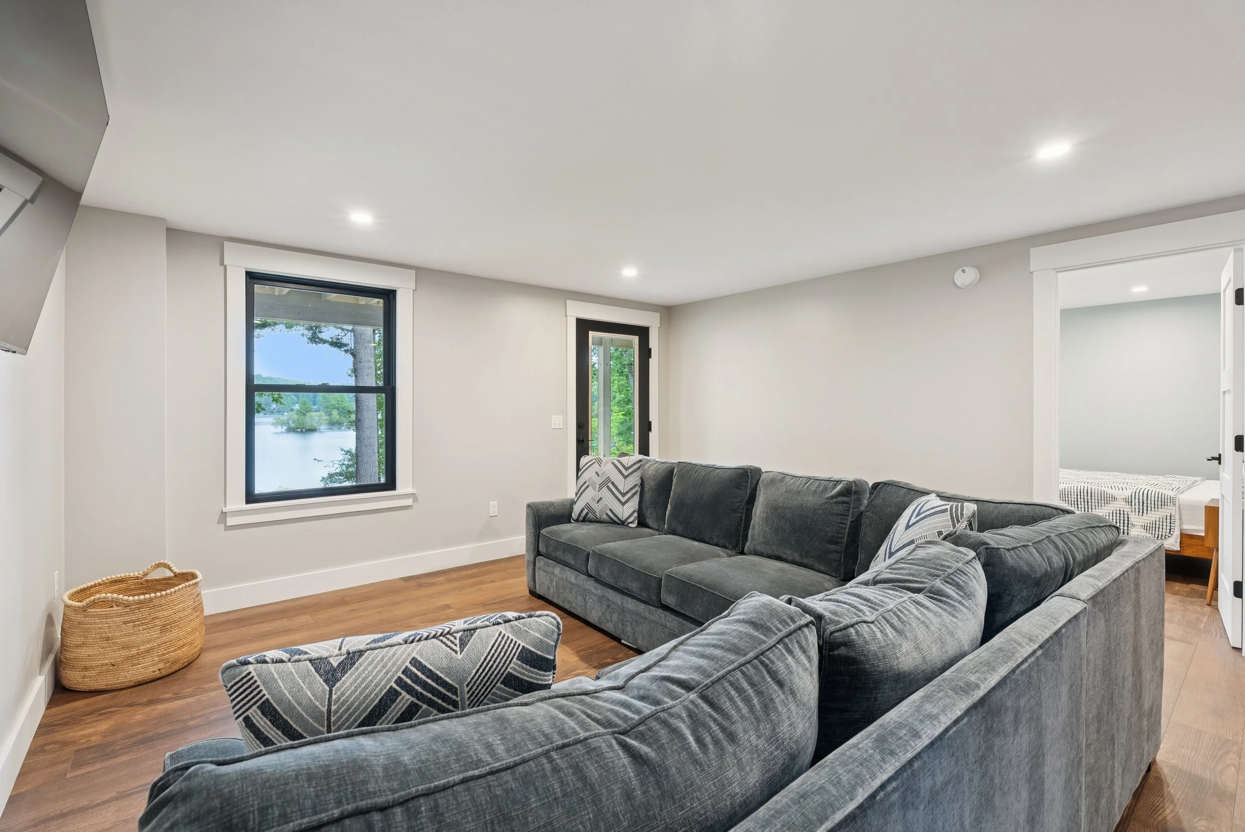 Living room with large gray sofas, a window showing trees and a lake outside, hardwood floors, and white walls.
