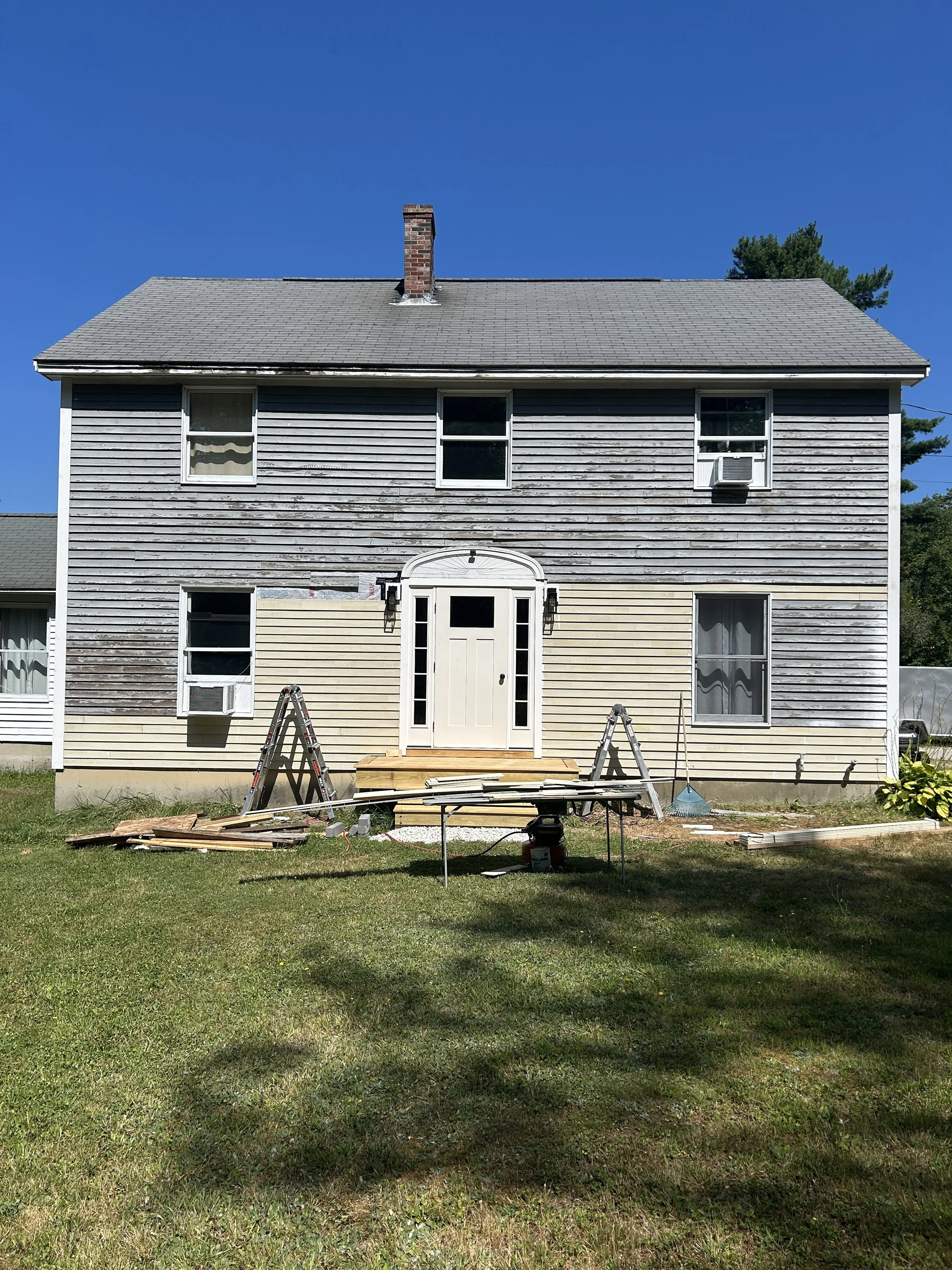 A two-story house with gray weathered wood siding on the upper level and yellow siding on the lower level, featuring a white front door with sidelight windows. The house has a pitched gray shingle roof and a brick chimney. There are multiple windows,