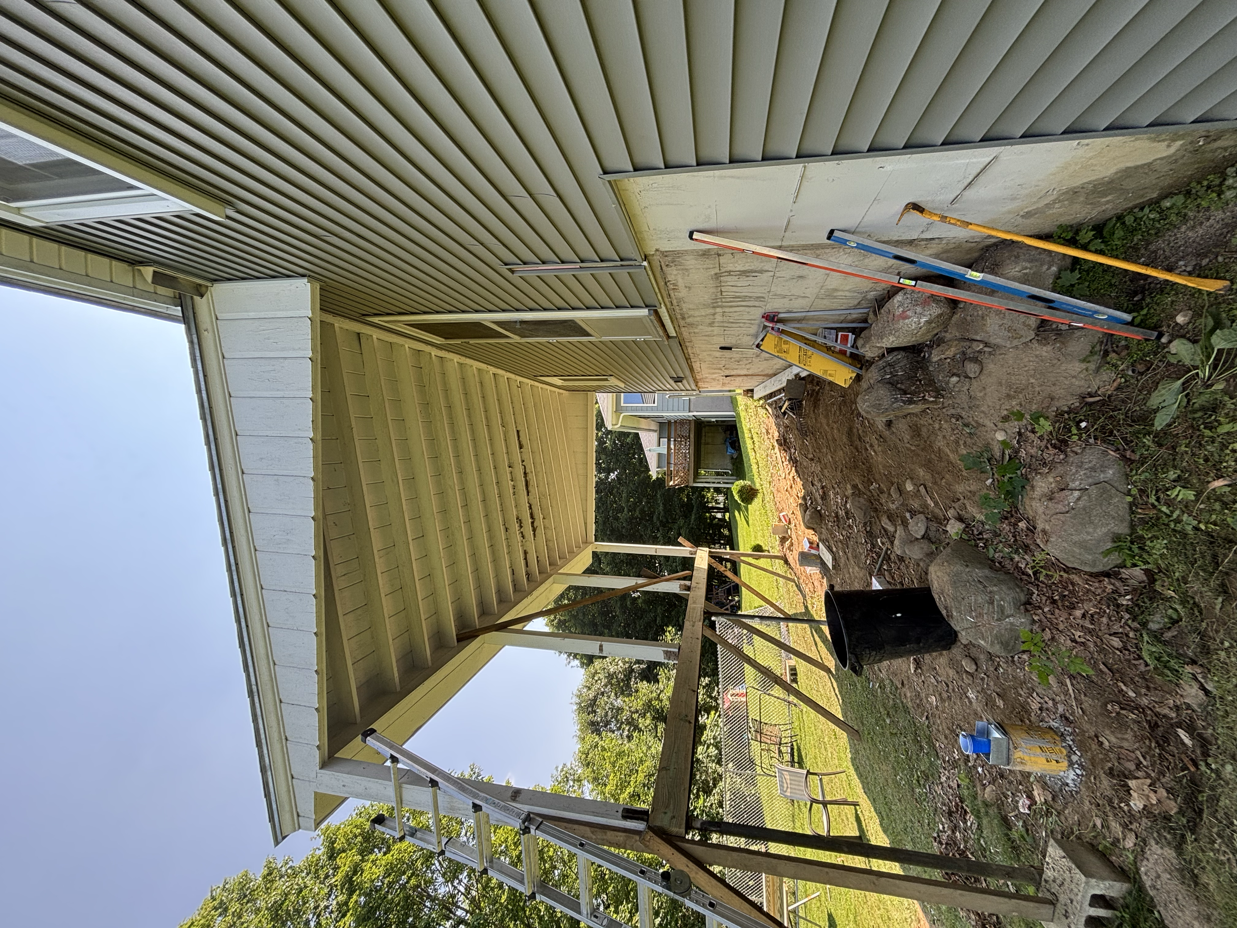 A yellow house with siding undergoing construction or renovation. The house has an overhanging roof, and there are construction tools and materials nearby, including a level, yellow caution tape, and a black container, with soil and rocks in the fore