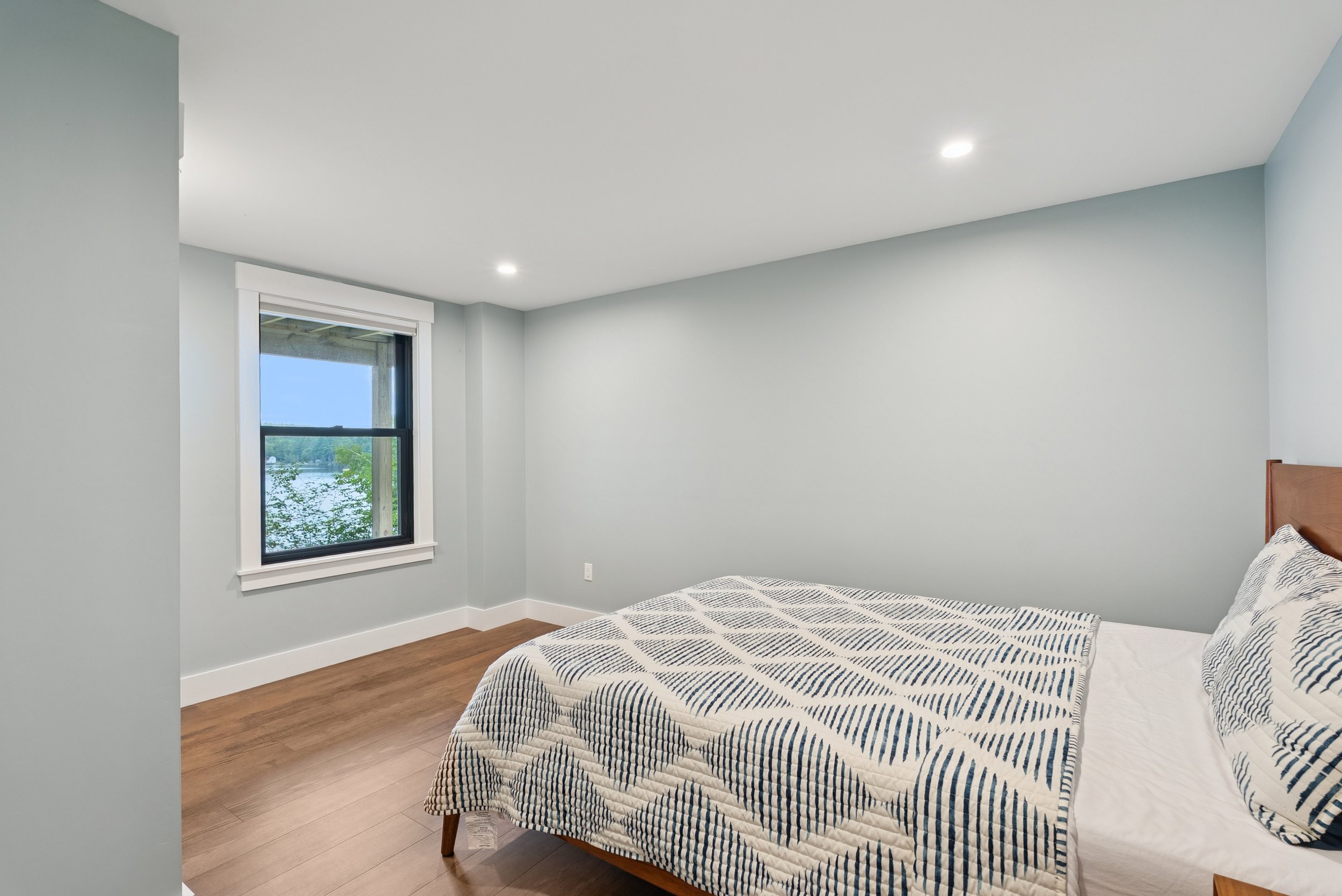 A bedroom with light blue walls, a window showing a view of water and trees, and a bed with a striped black and white quilt.