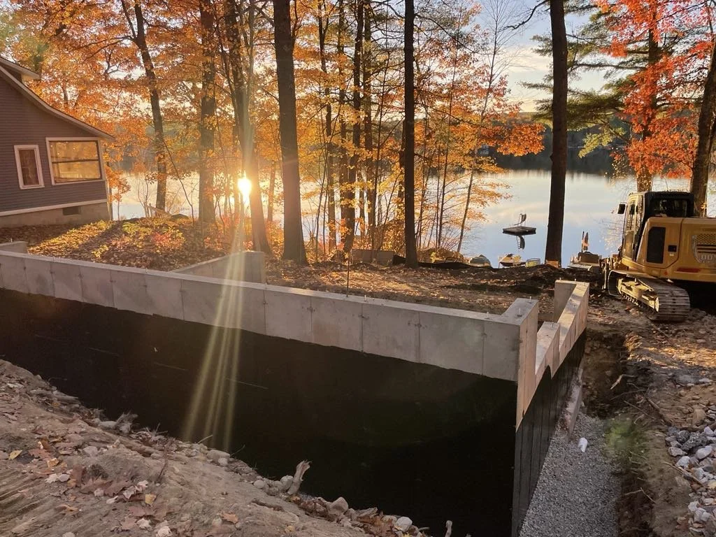 Construction site by a lake during fall, with a partially built retaining wall, a yellow excavator, a house on the left, and trees with orange and yellow leaves, as the sun sets over the water.