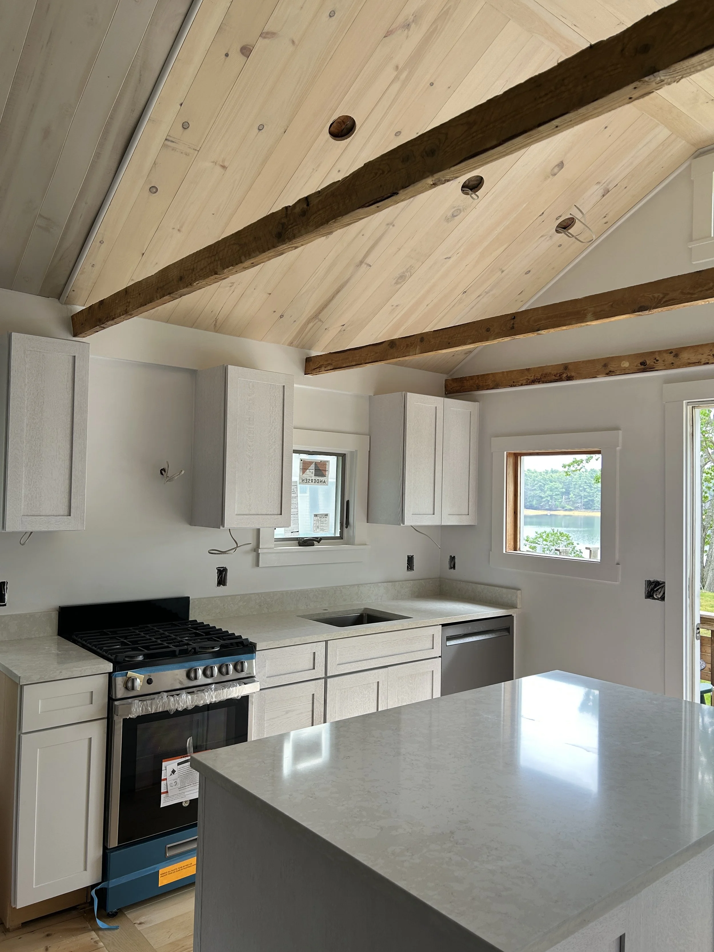 Modern kitchen with white cabinets, a light-colored countertop, a stove, and a large window with a view of a lake and trees outside. The ceiling has wooden beams and light wood paneling.
