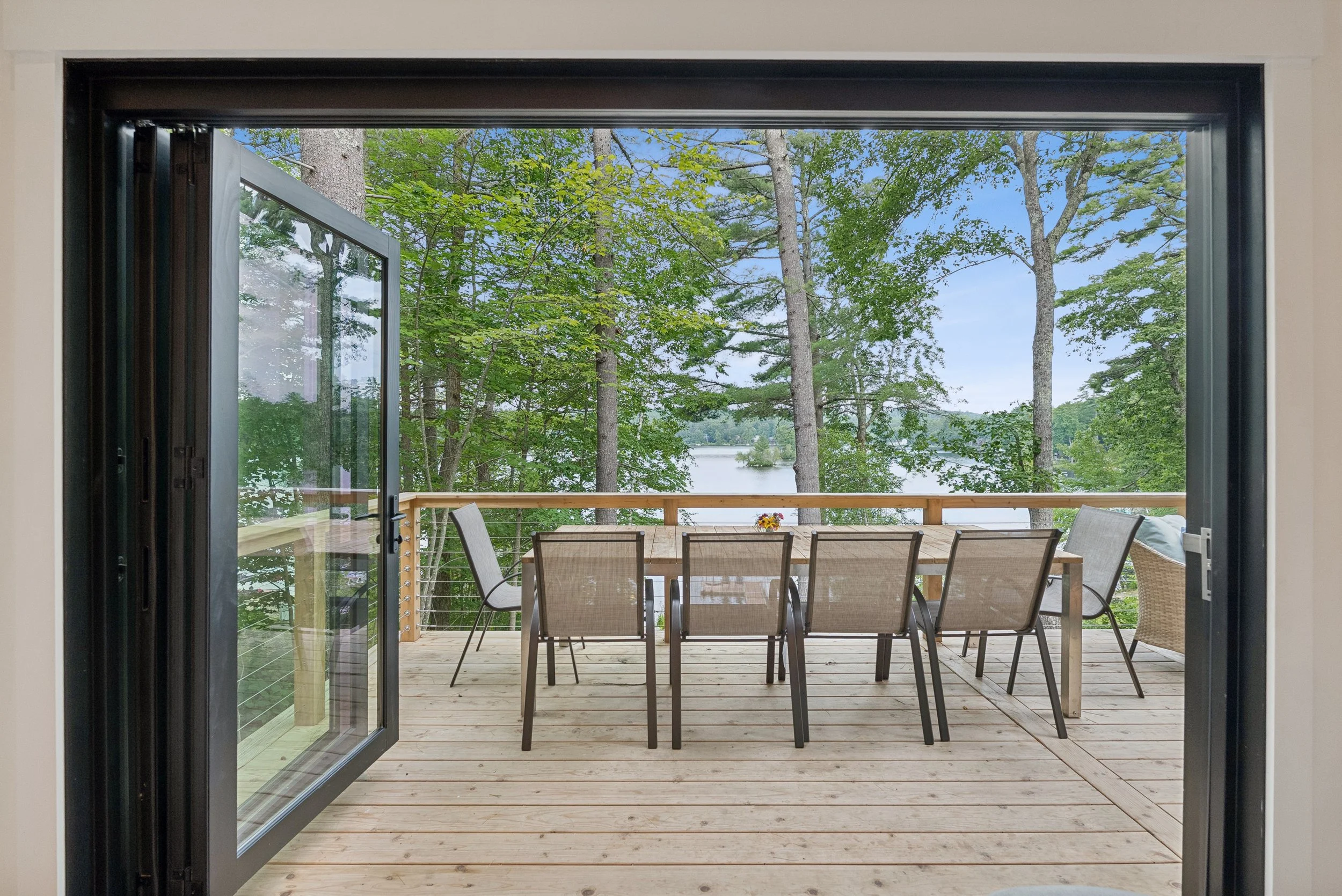 A view of a wooden deck with a dining table and chairs, opening to a lush green forest with a lake visible in the background.