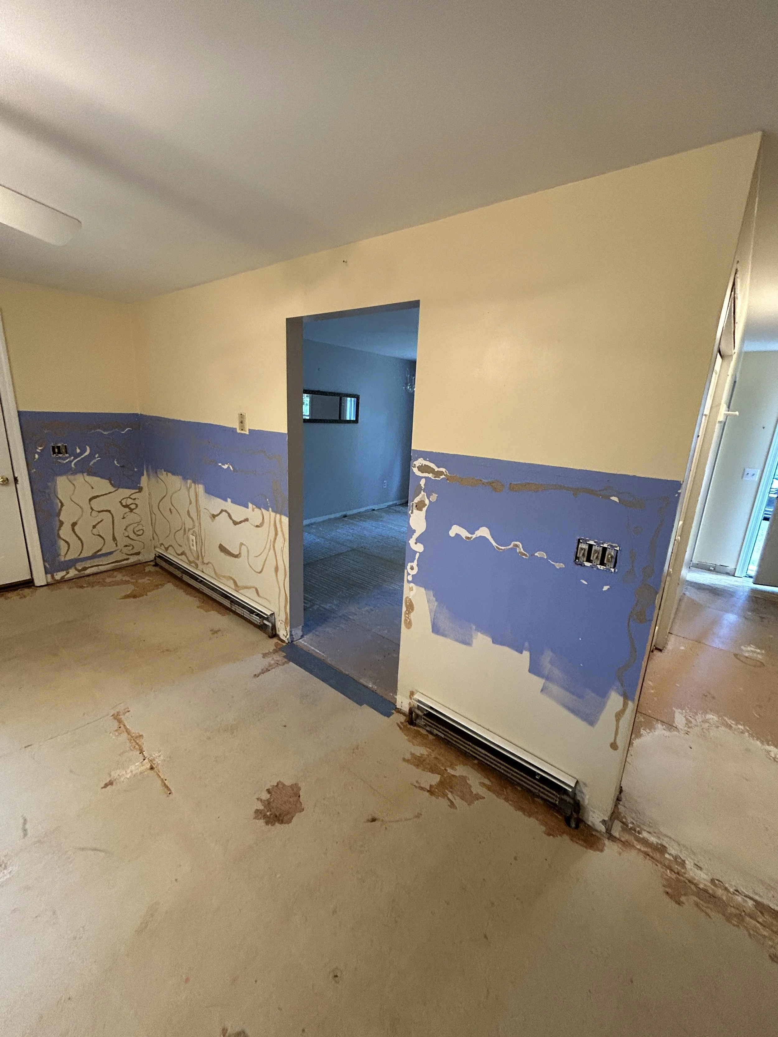 Living room during renovation with walls partially painted blue and yellow, exposed wall sockets, and a damaged floor with paint splatters.