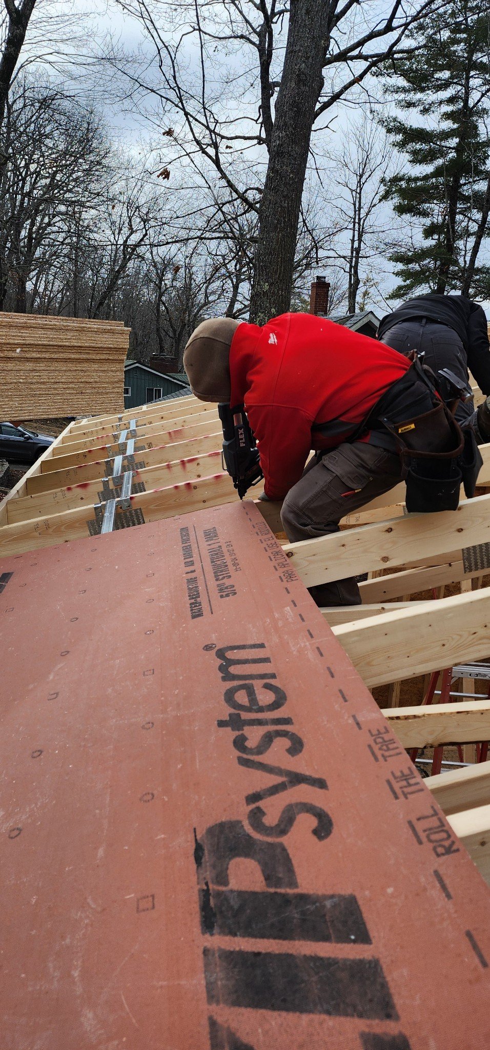 A construction worker in a red jacket and beige hat is working on wooden framing on an outdoor construction site. There are trees and a house in the background.