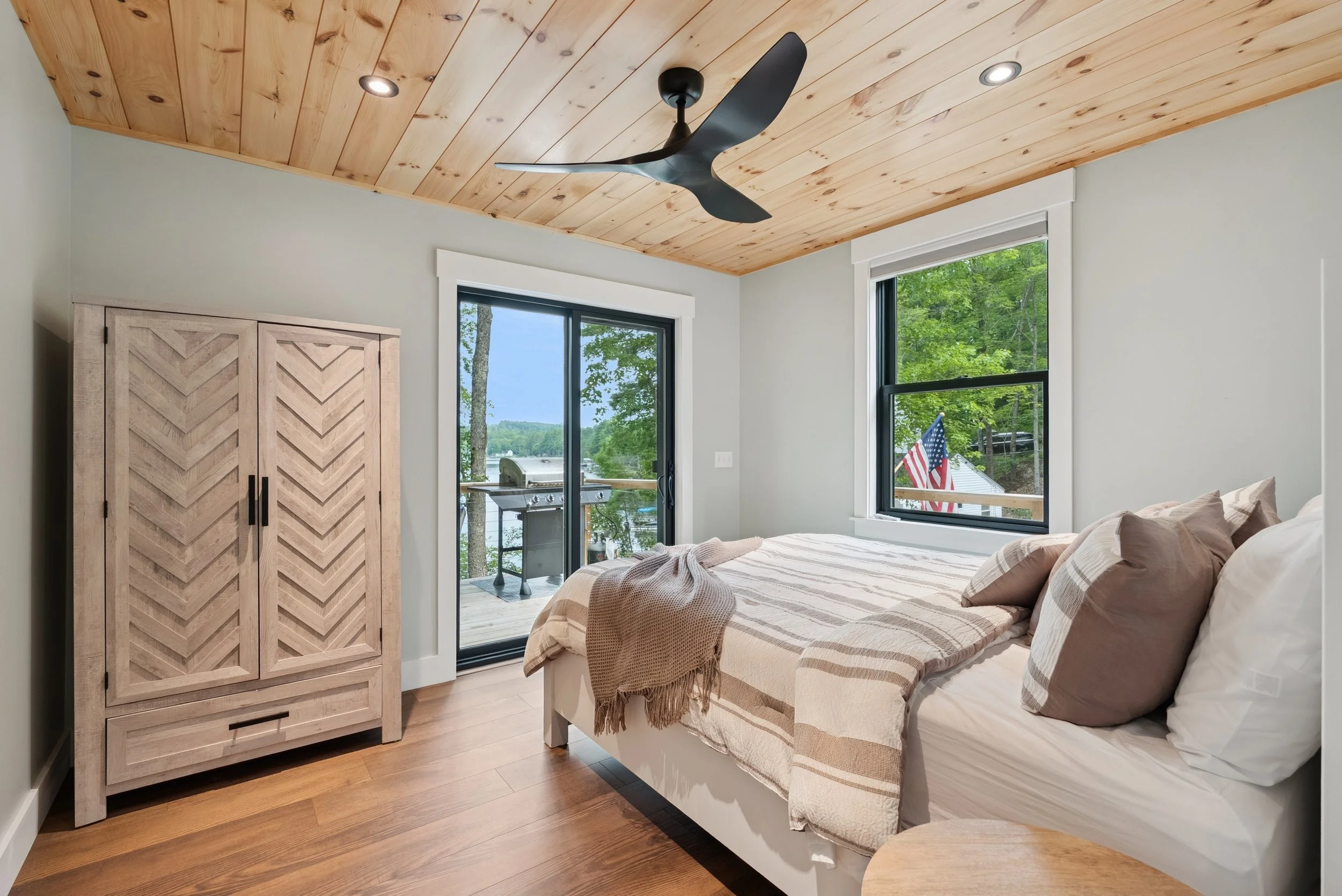 Bedroom with wooden ceiling and light-colored walls, large windows showing trees, a sliding glass door leading to a deck with a grill, and a bed with striped bedding and pillows.