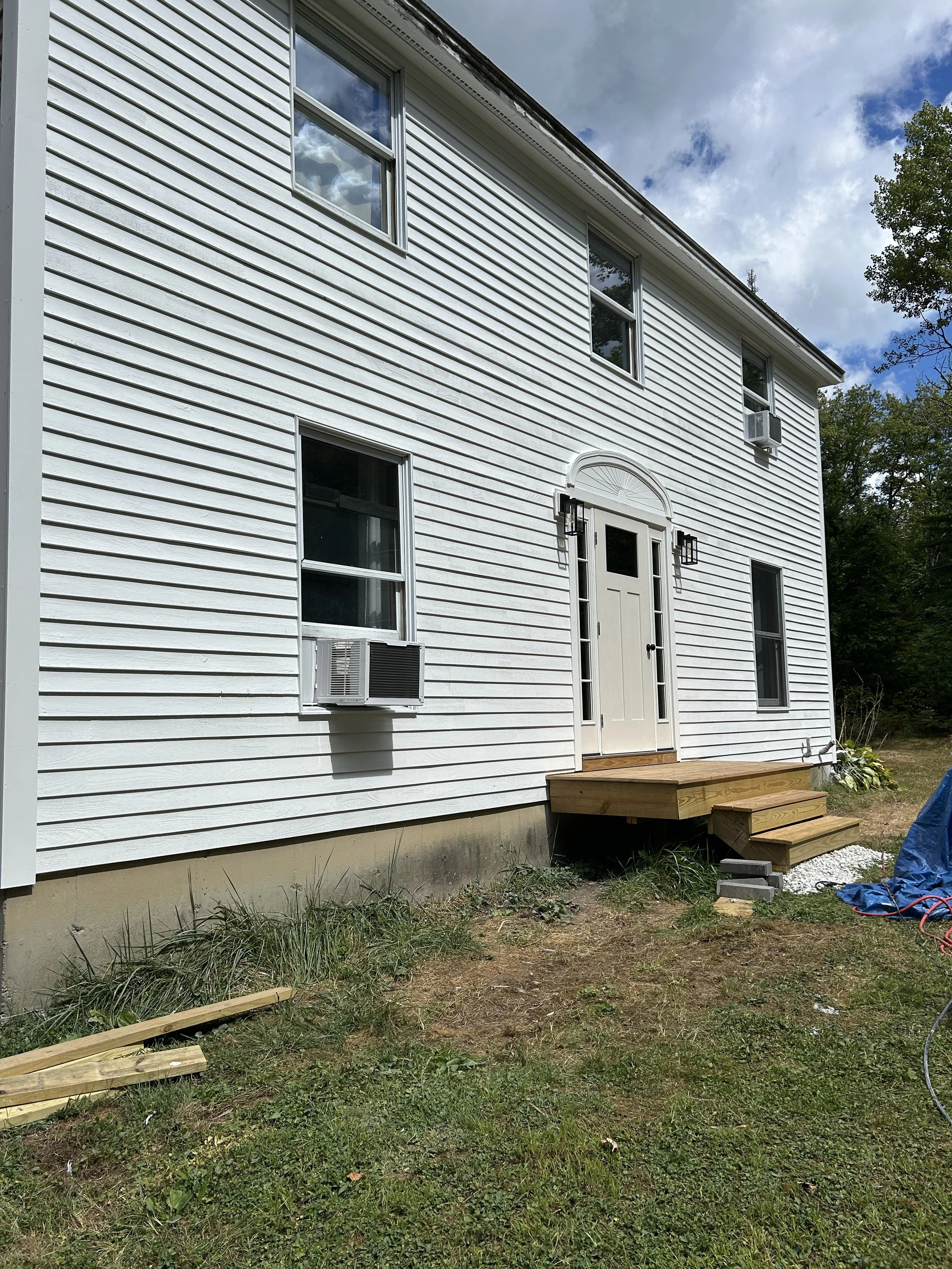 Back of a white, two-story house with horizontal siding, three visible windows, and a front door. There's a small wooden staircase leading to the door, with construction materials and tools on the ground nearby.