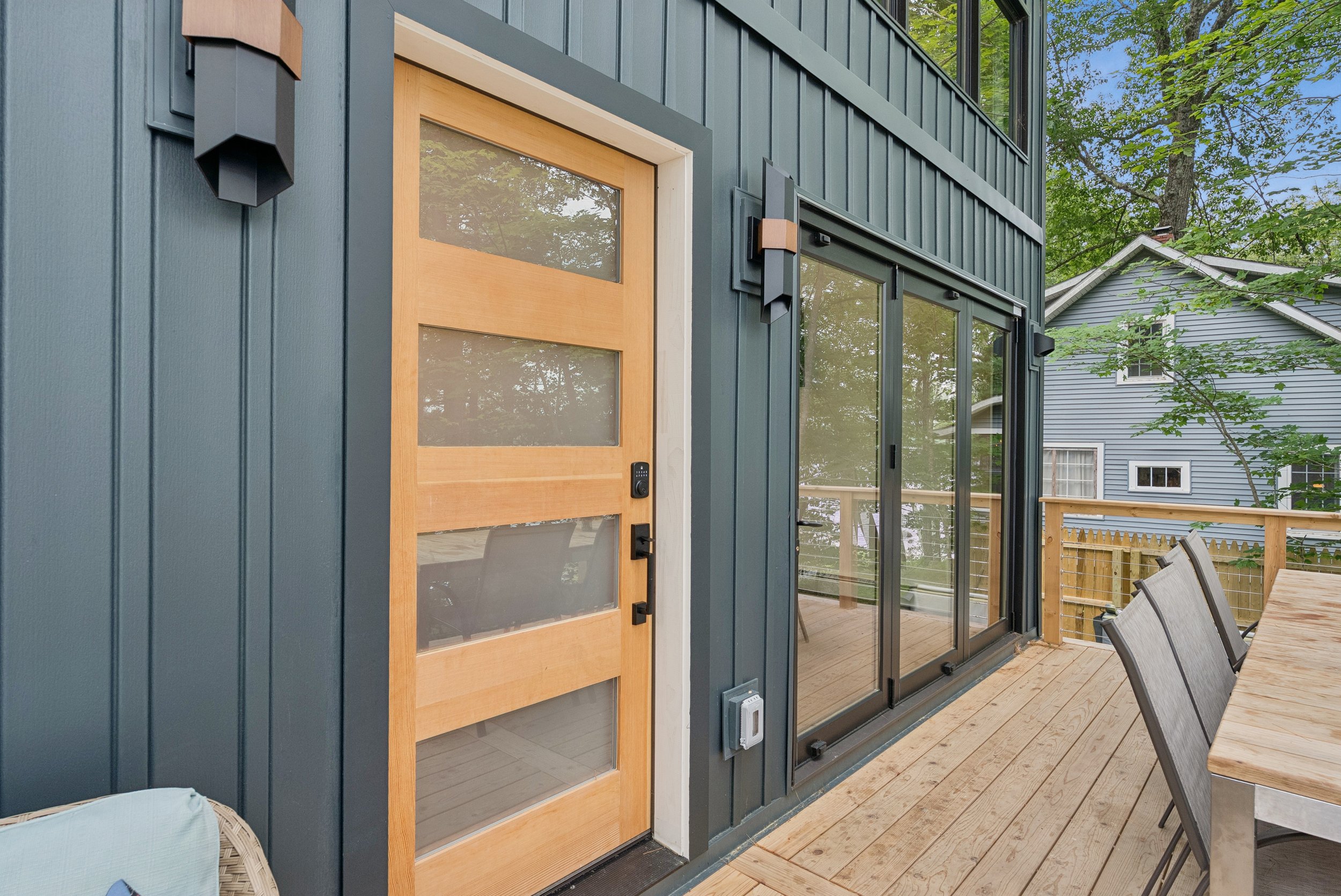 Exterior view of a modern house showing a light wooden front door with glass panels, dark green vertical siding, large sliding glass door leading to a deck, outdoor furniture, and trees in the background.
