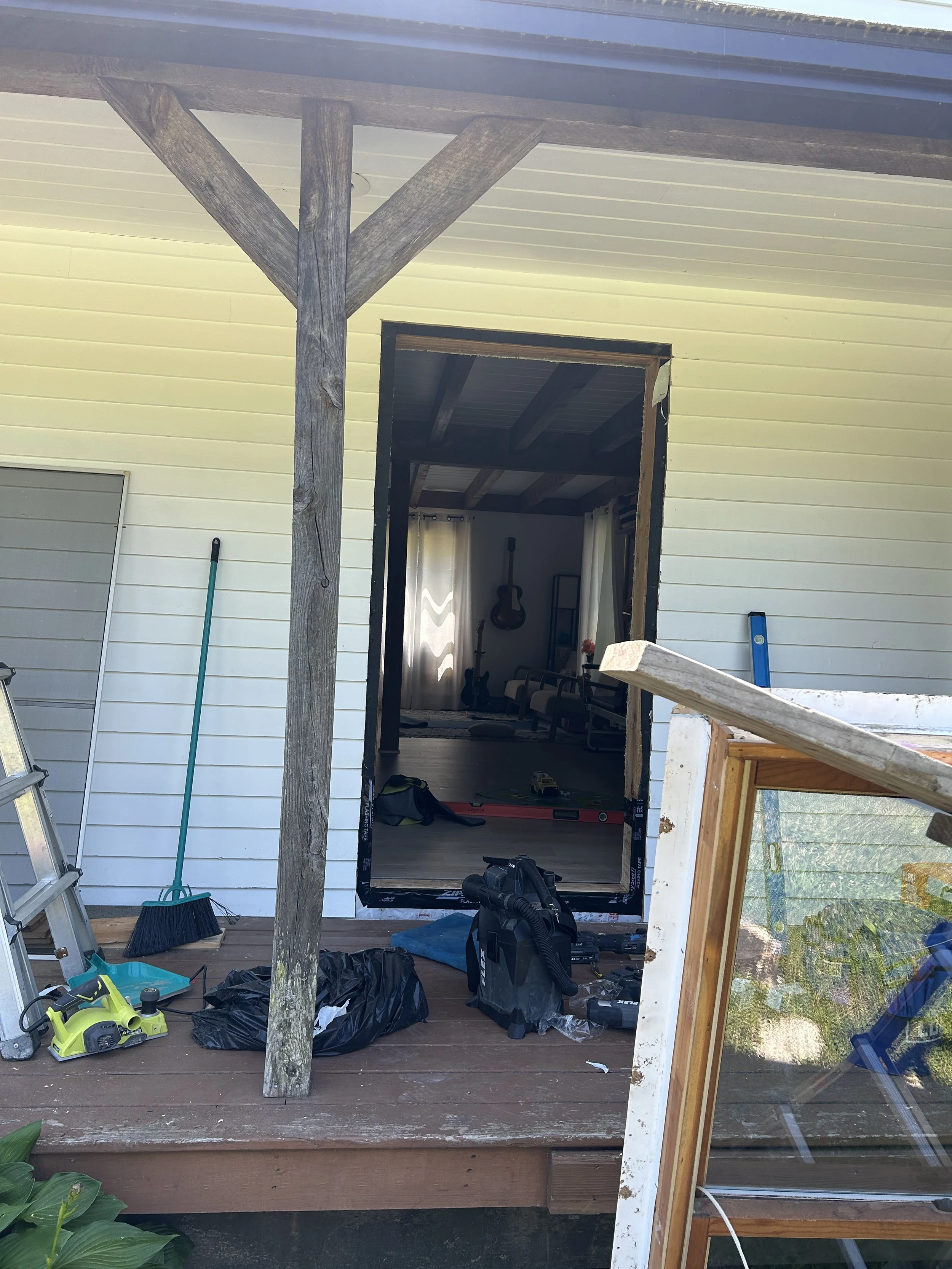 Image of a house porch area under construction or renovation, with tools and materials scattered around including a green powered tool, a broom, a ladder, a large black trash bag, and a window frame leaning against the porch. The porch has a wooden f