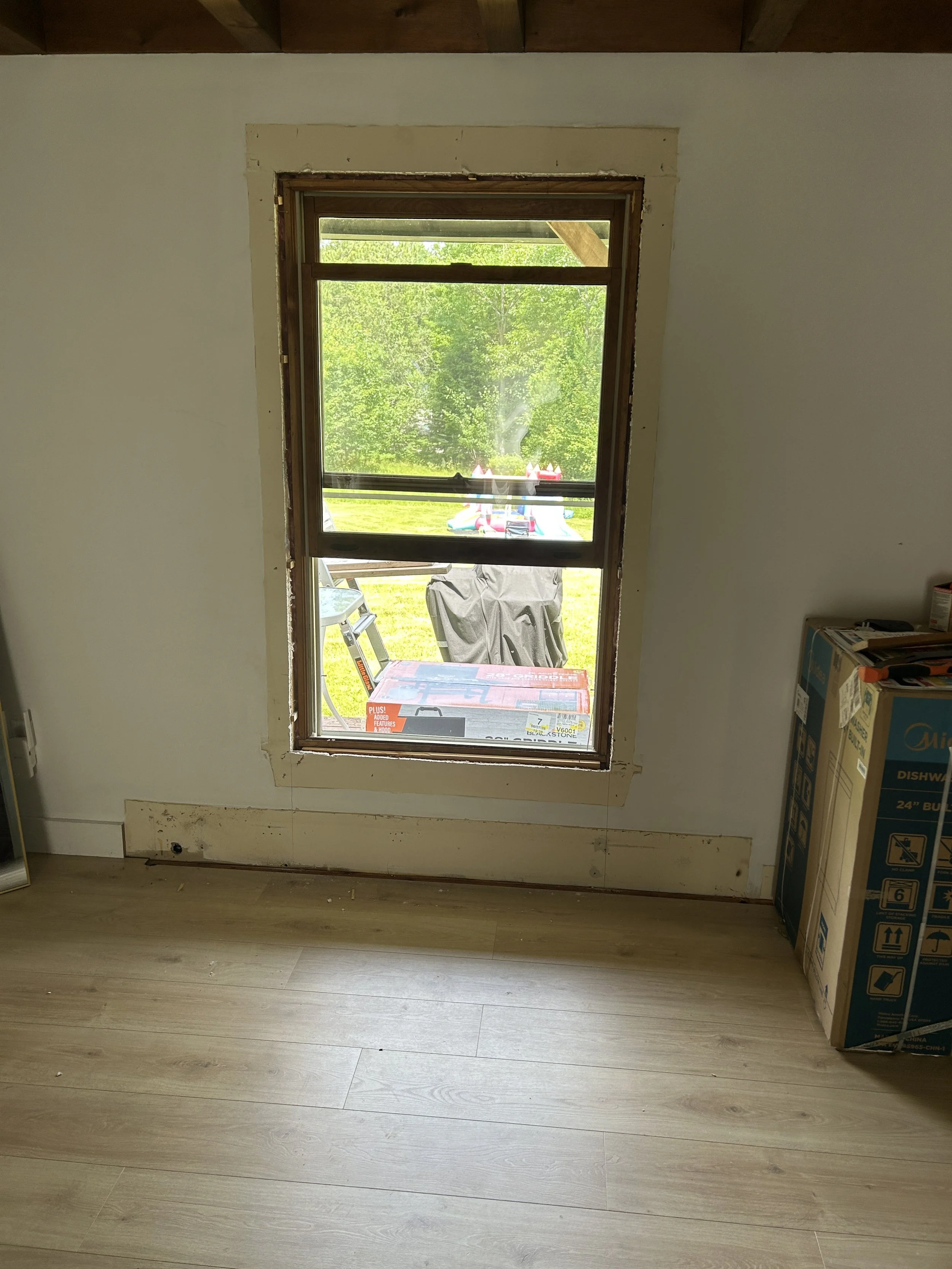 A room with a window looking outside at green trees and a playground with a bounce house. The wall around the window is unfinished, and there is a cardboard box of a dishwasher on the floor to the right.