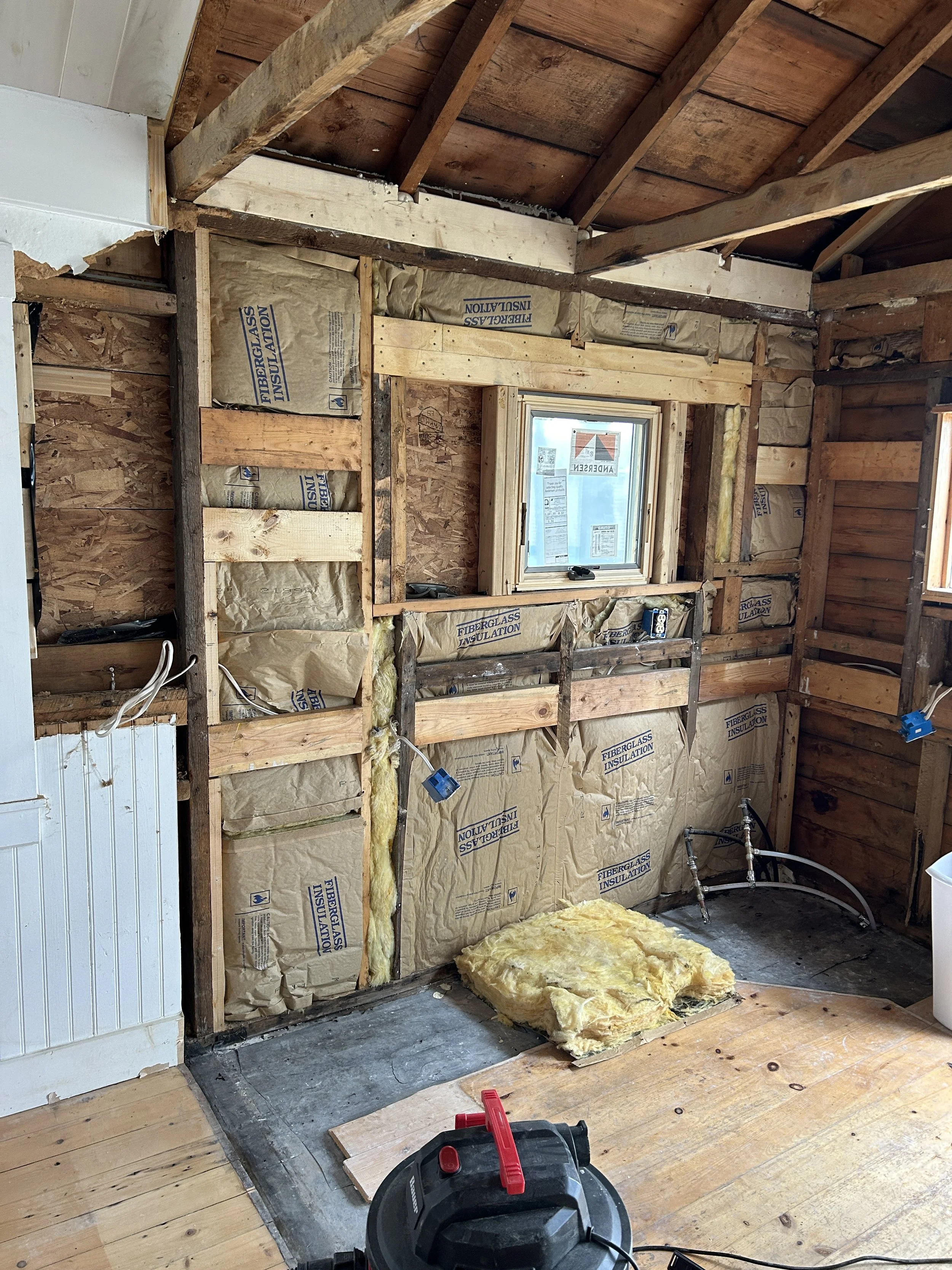 A room under renovation with exposed wooden framing and fiberglass insulation in the walls. There is a small window and construction tools on the floor, with some yellow insulation on the ground.