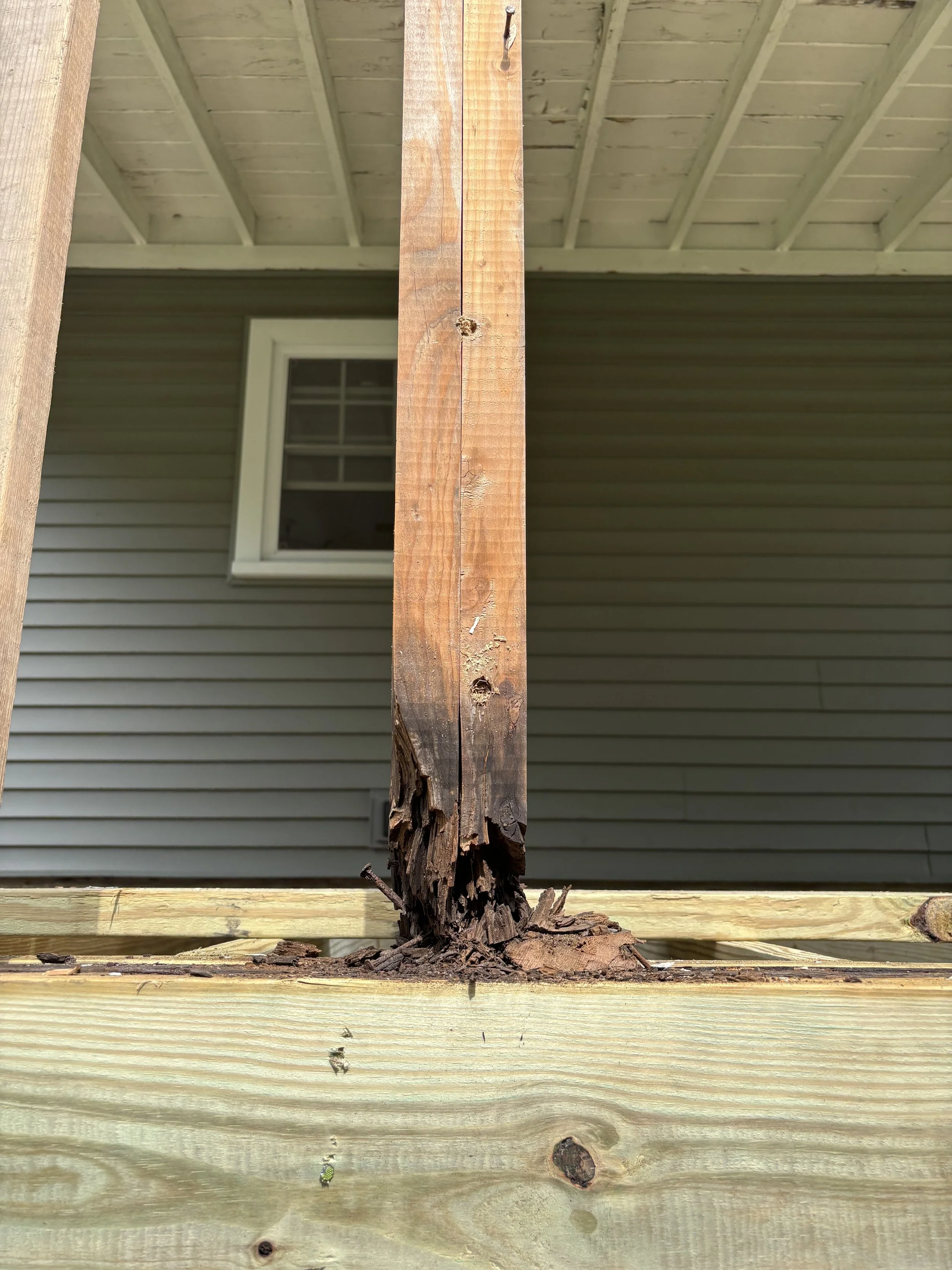 A close-up of a damaged wooden post supporting a deck, with worn and splintered wood at the bottom. A house with vinyl siding and a window are in the background.