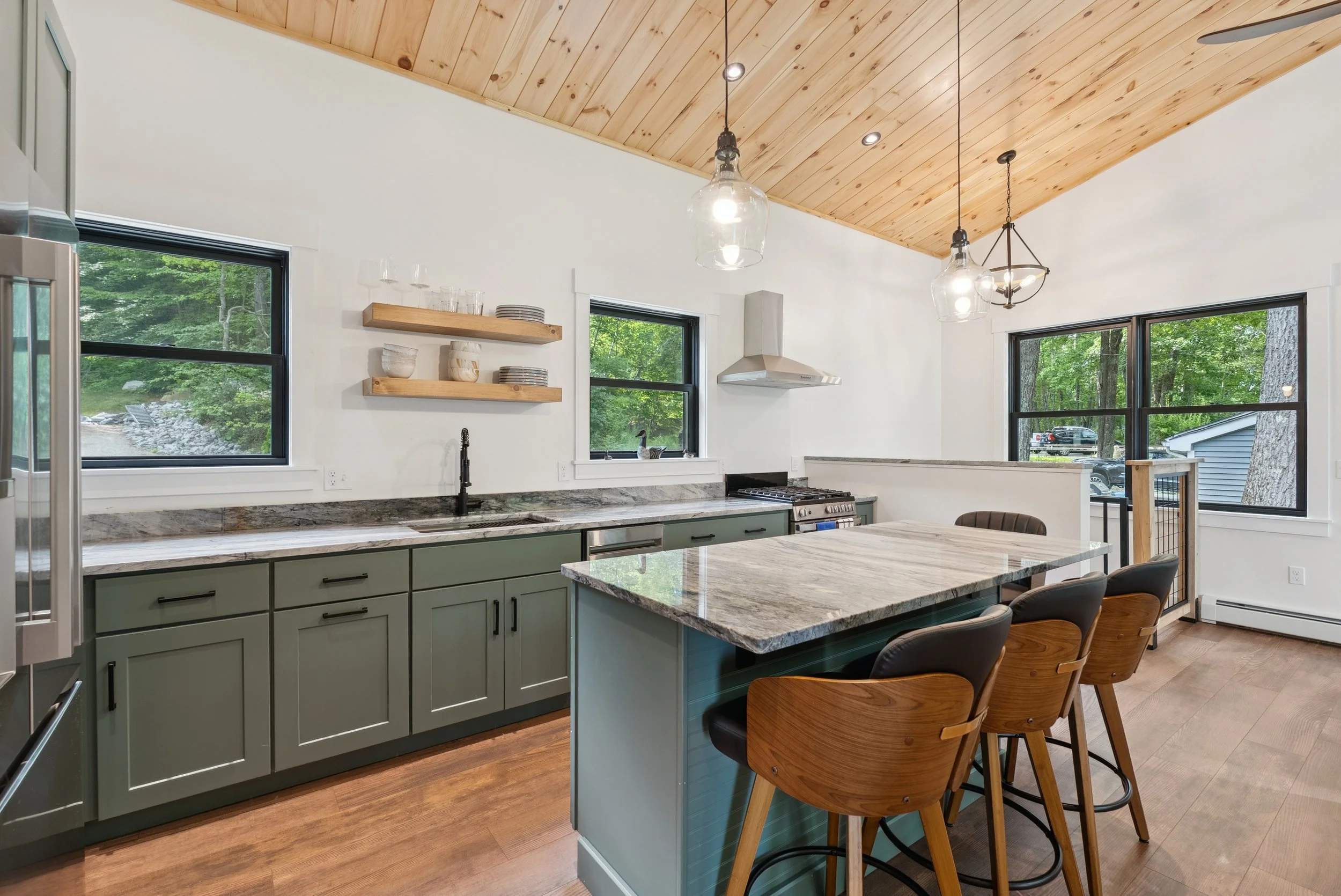 Modern kitchen with green cabinets, granite countertop, wooden bar stools, natural wood vaulted ceiling, and large windows showing trees outside.
