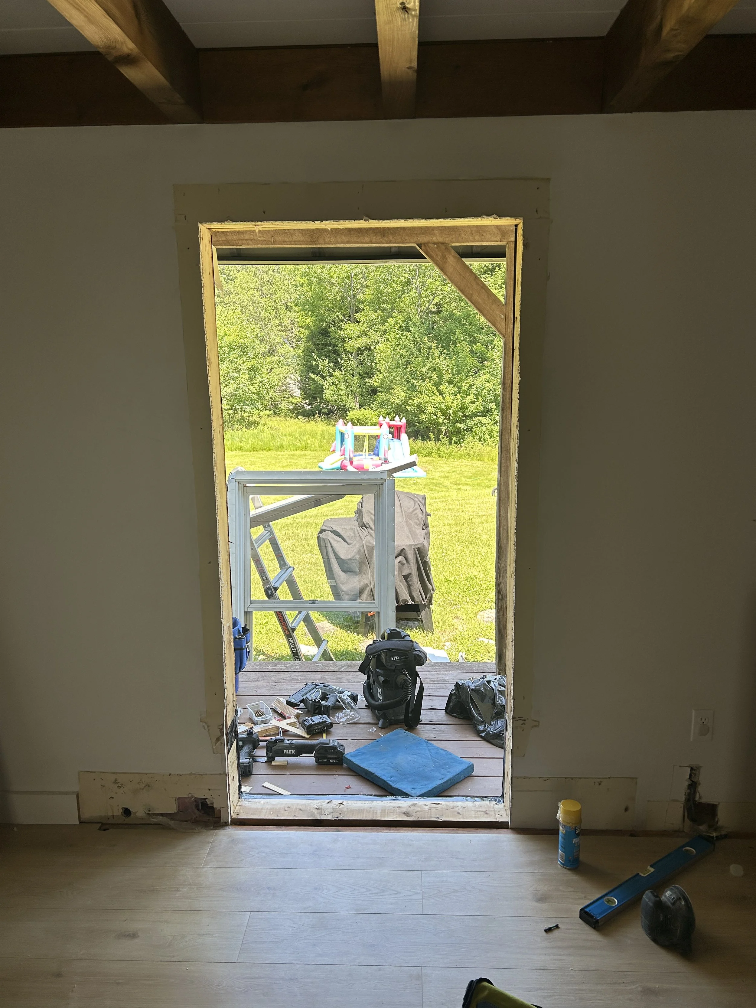 View through a doorless opening in a room under renovation, leading to a backyard with a children's inflatable bounce house and green trees in the background. Construction tools and supplies are on the floor and outside on the porch.