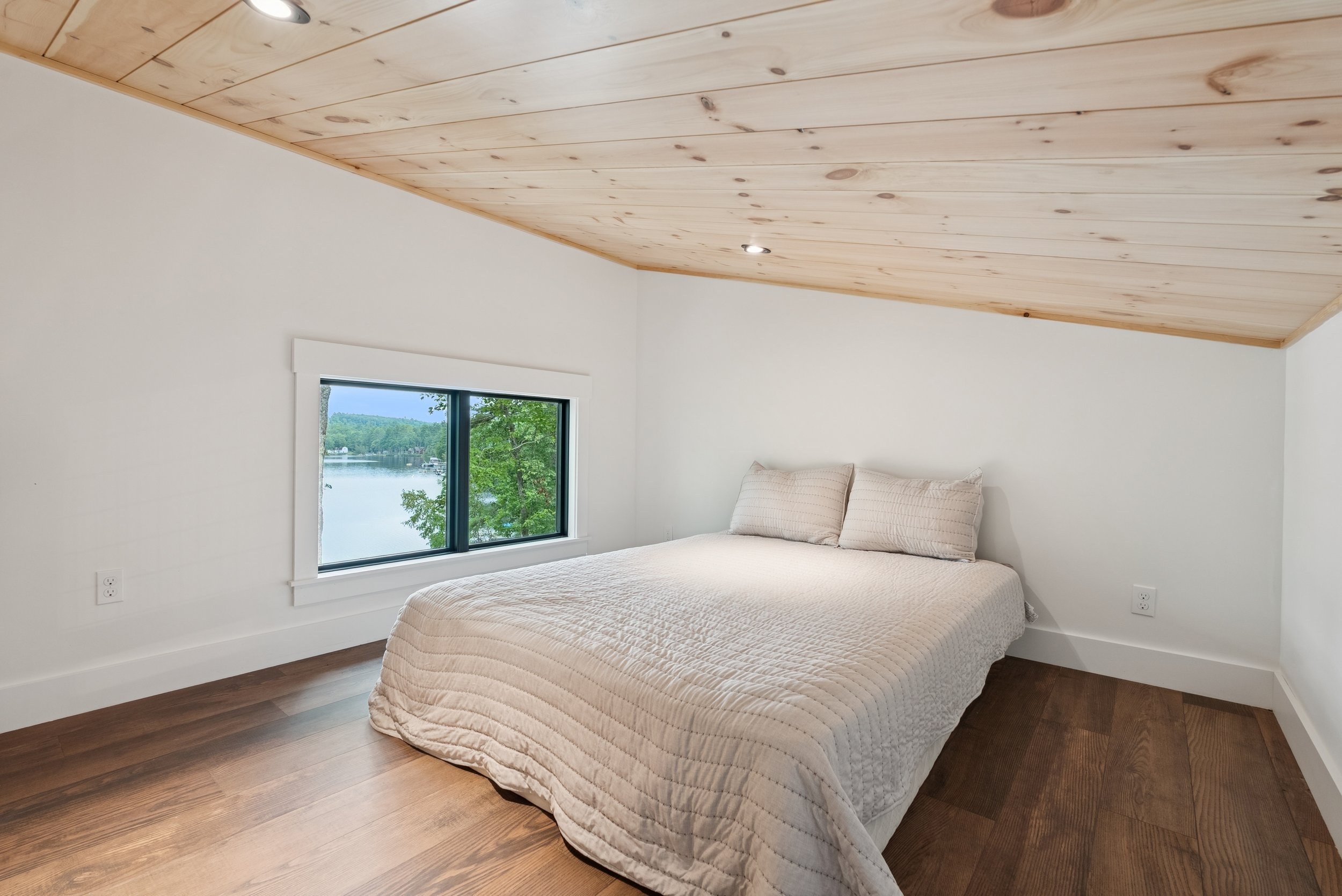 A bedroom with a vaulted ceiling covered in light wood panels, a window showing a scenic view of a lake and trees, a bed with a beige quilt and pillows, and hardwood flooring.