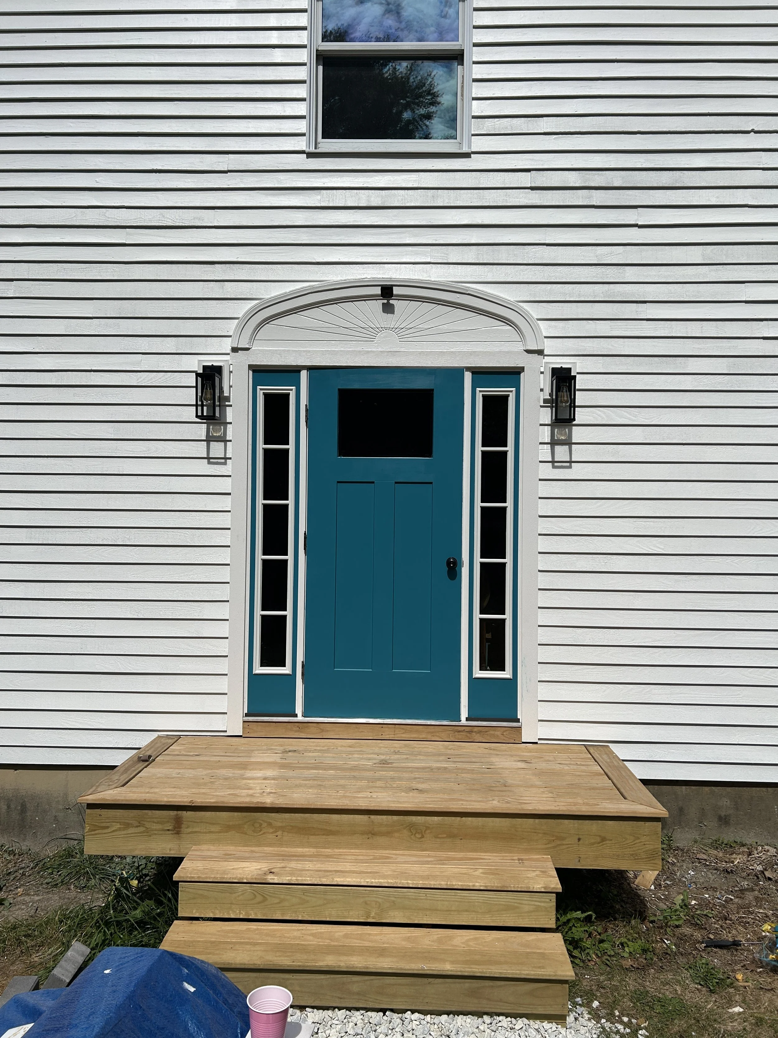 The image shows a new wooden deck leading up to a house with white horizontal siding. The house has a bright blue door with glass sidelights and a transom window above. There are two black wall-mounted lantern-style lights on either side of the door,