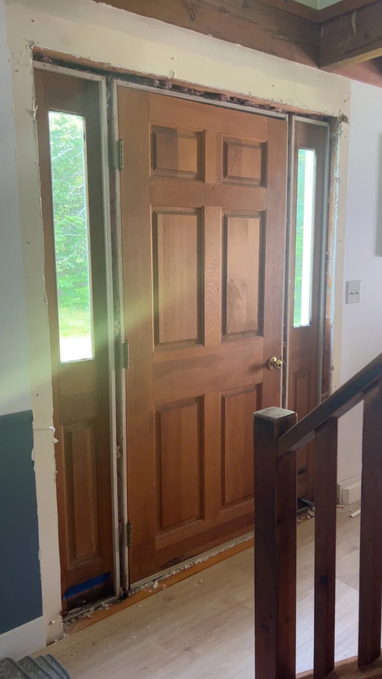 Wooden front door with glass side panels under construction, with visible framing and interior wall damage, next to a staircase railing.