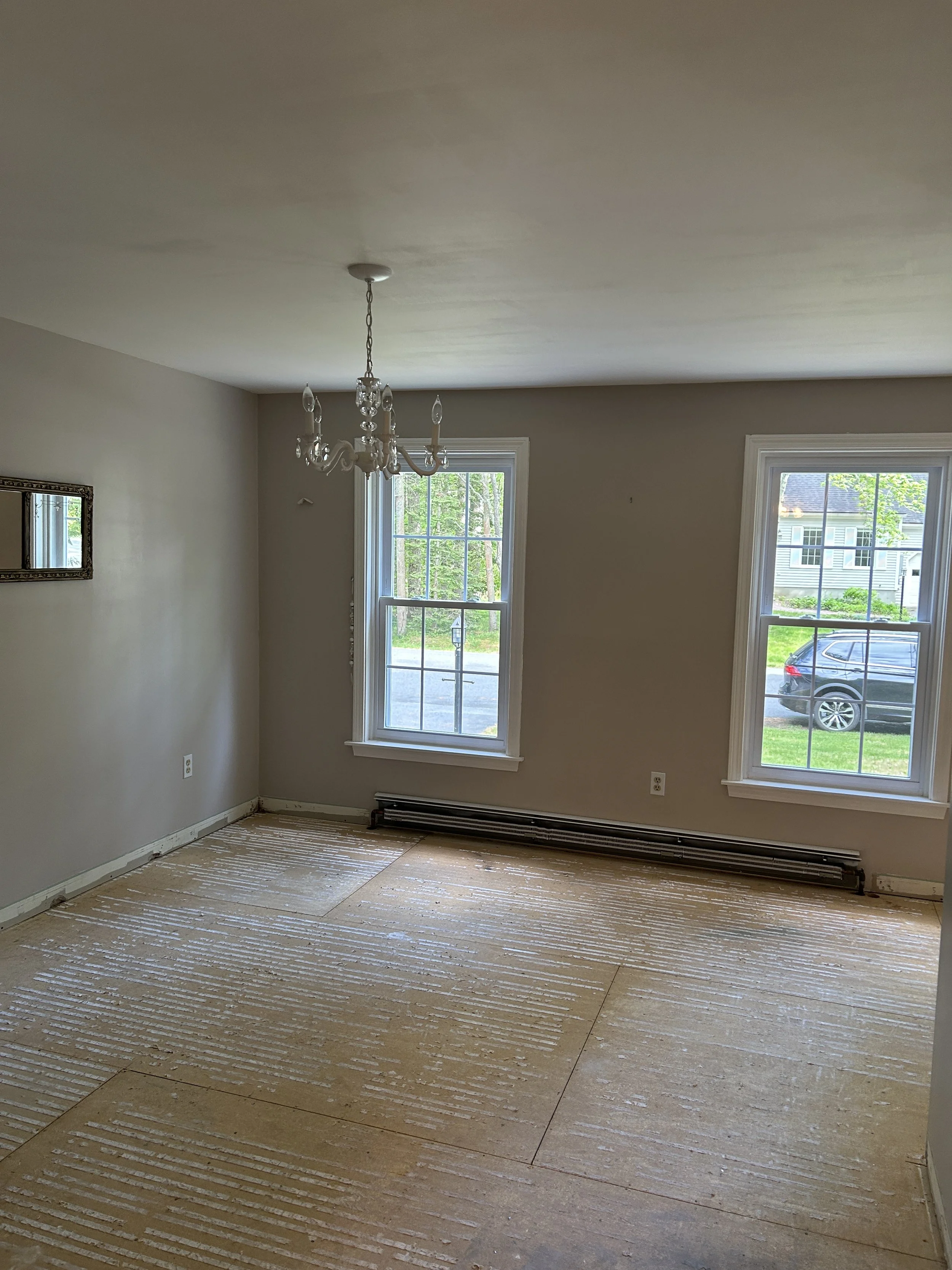 Room with large windows, chandelier, and exposed floor with broken flooring material.