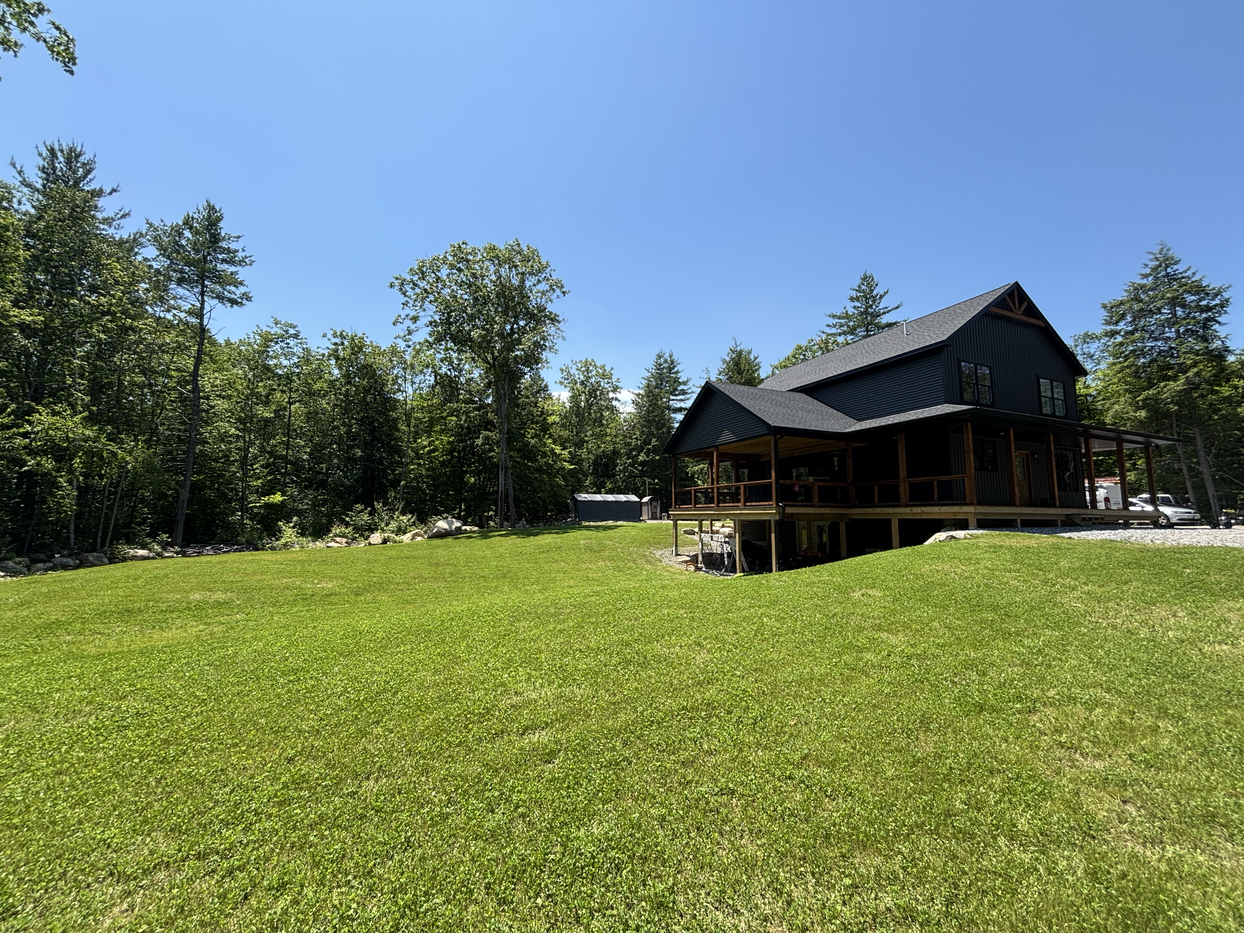 A house with black siding and large windows, surrounded by green trees and a well-maintained lawn, under a clear blue sky.