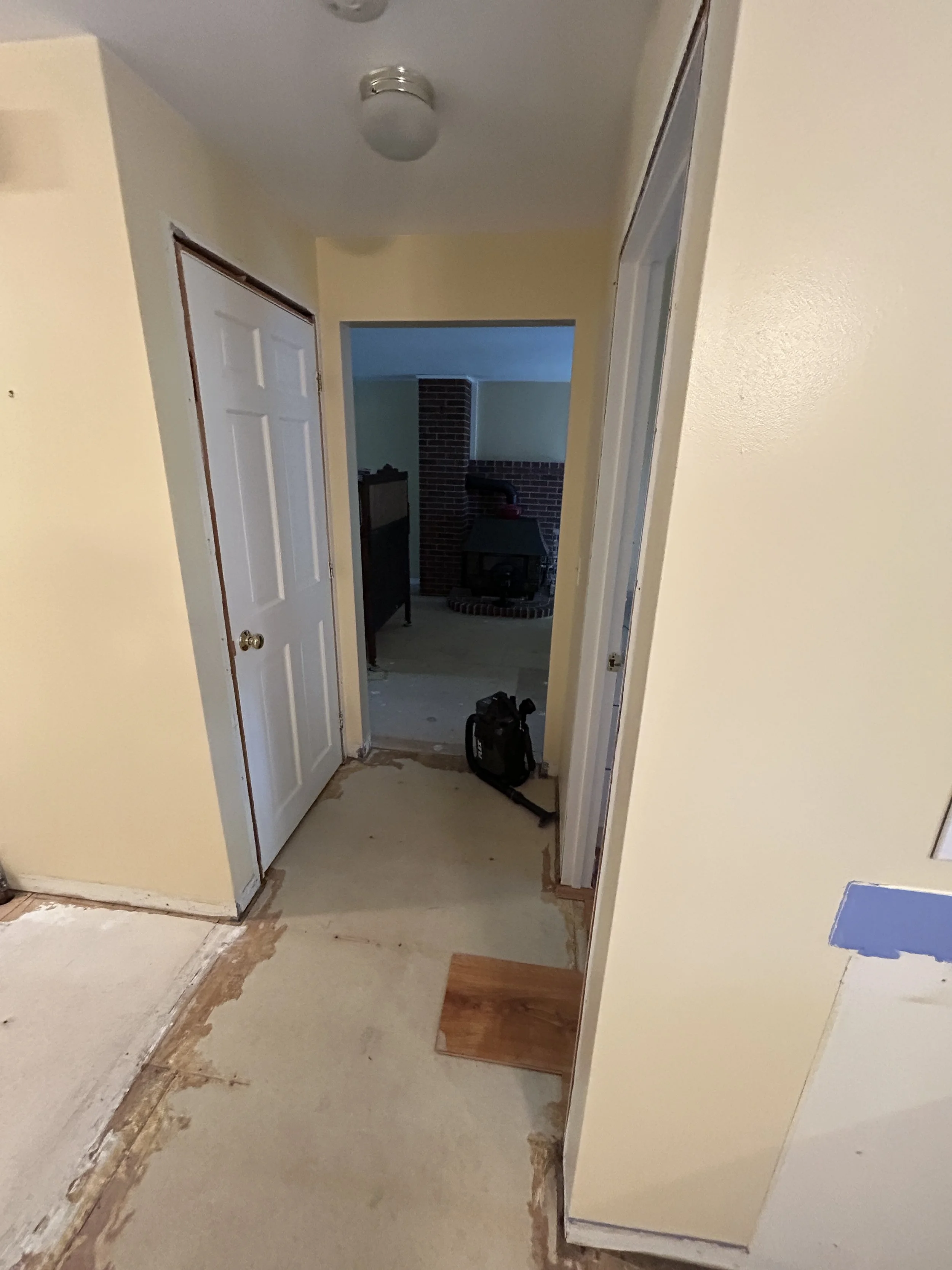 Interior of a house under renovation with yellow walls, a white door, a hallway leading to a brick fireplace, and a black backpack on the floor.