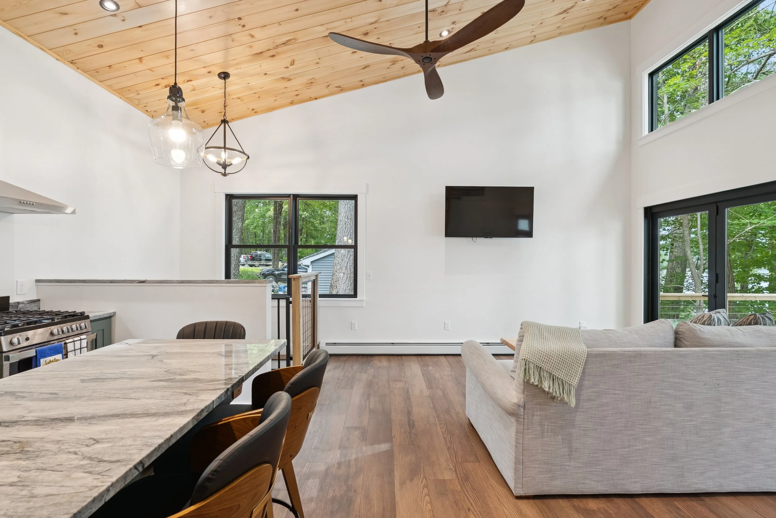 Interior view of a modern living room with a wooden vaulted ceiling, ceiling fan, large windows showing a green outdoor scene, white walls, a flat-screen TV, a dining table with chairs, and a beige sofa with a blanket.