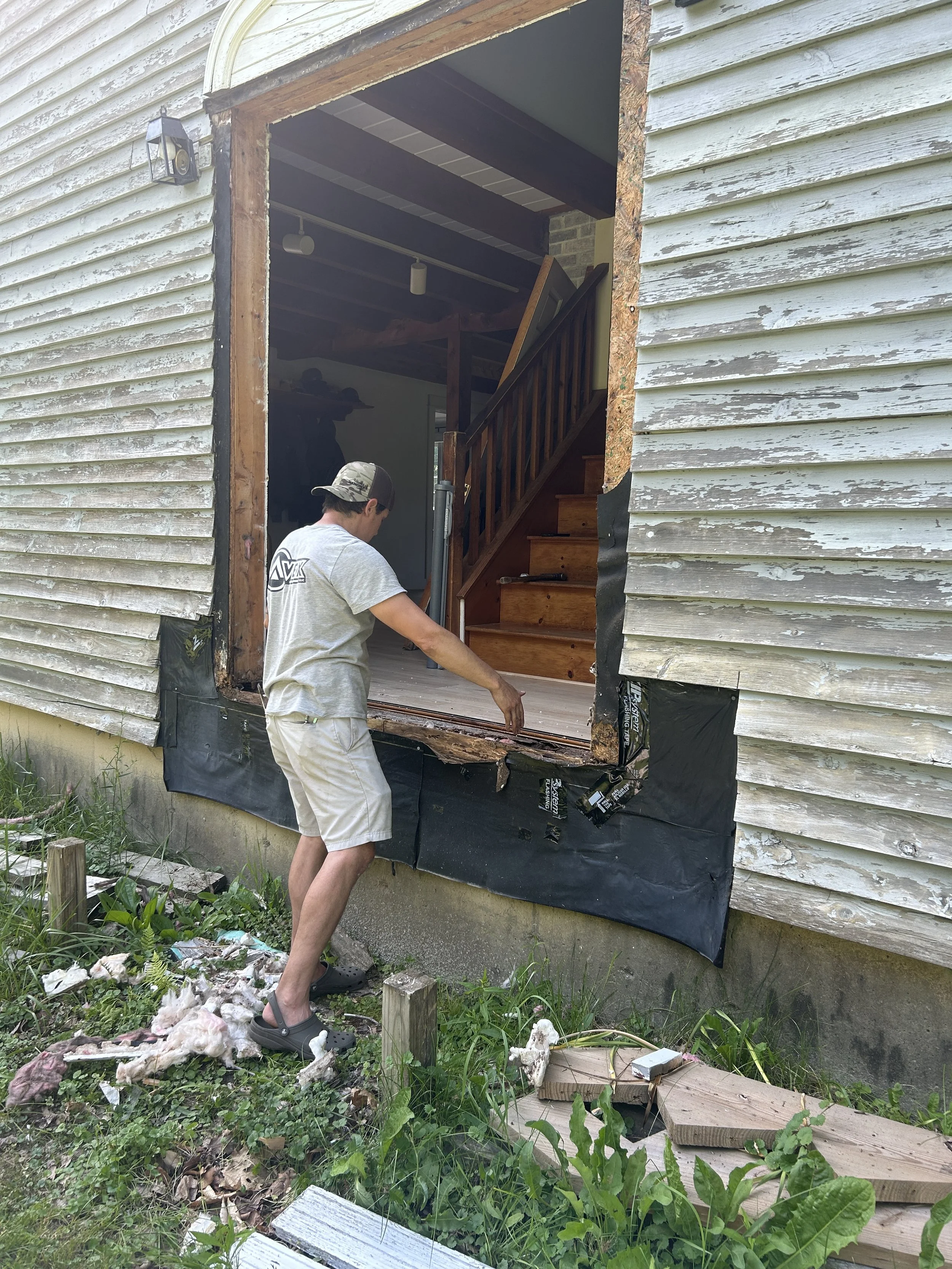 A man working on removing a window from the exterior of a house, which has peeling white paint and black water-resistant membrane at the bottom. Debris, insulation, and wood scraps are scattered on the ground outside.