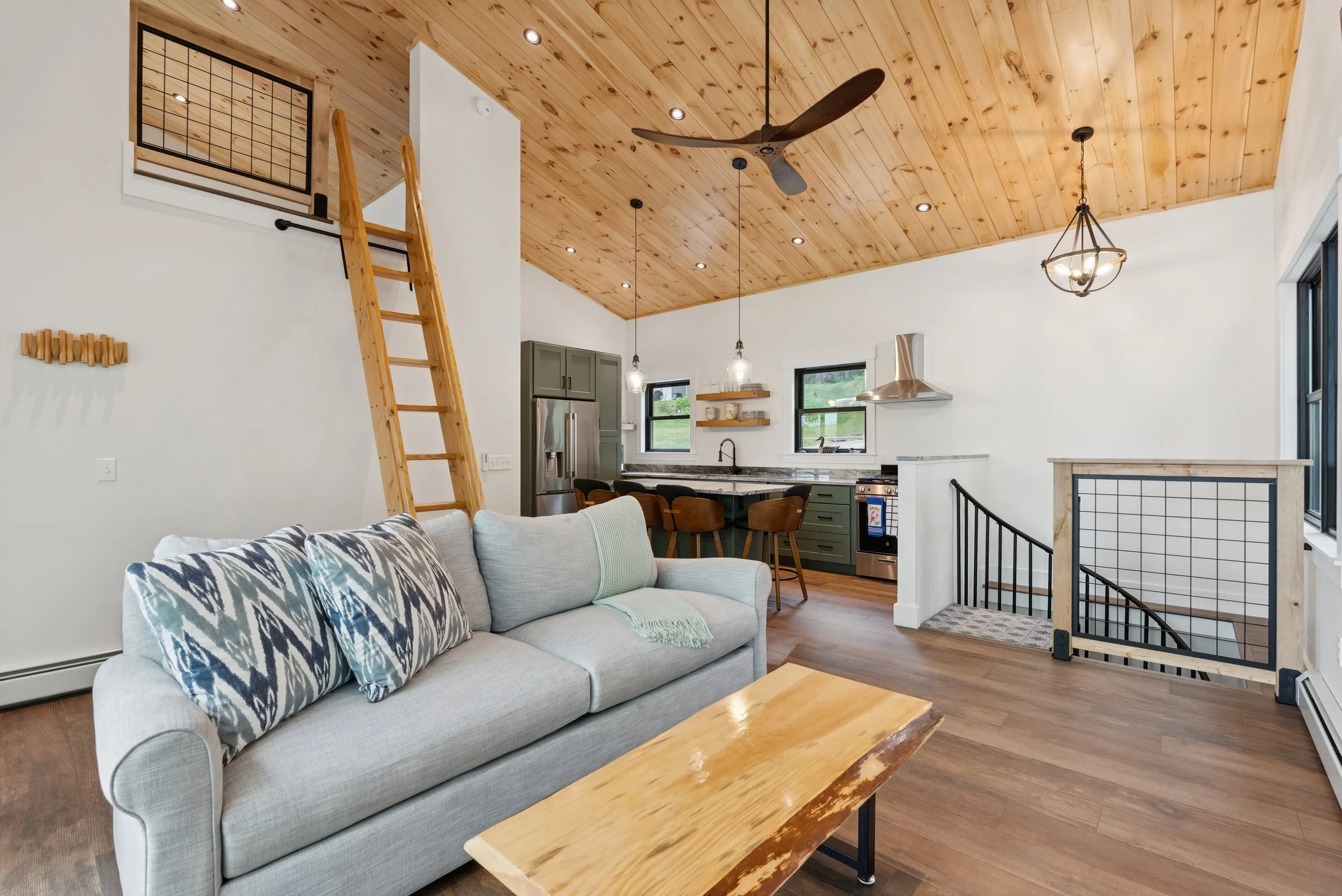 Open-concept living room and kitchen with wood-paneled ceiling, ceiling fan, pendant lights, large windows, gray sofa with patterned pillows, wooden coffee table, kitchen cabinets, stainless steel refrigerator, black dining chairs, and staircase with