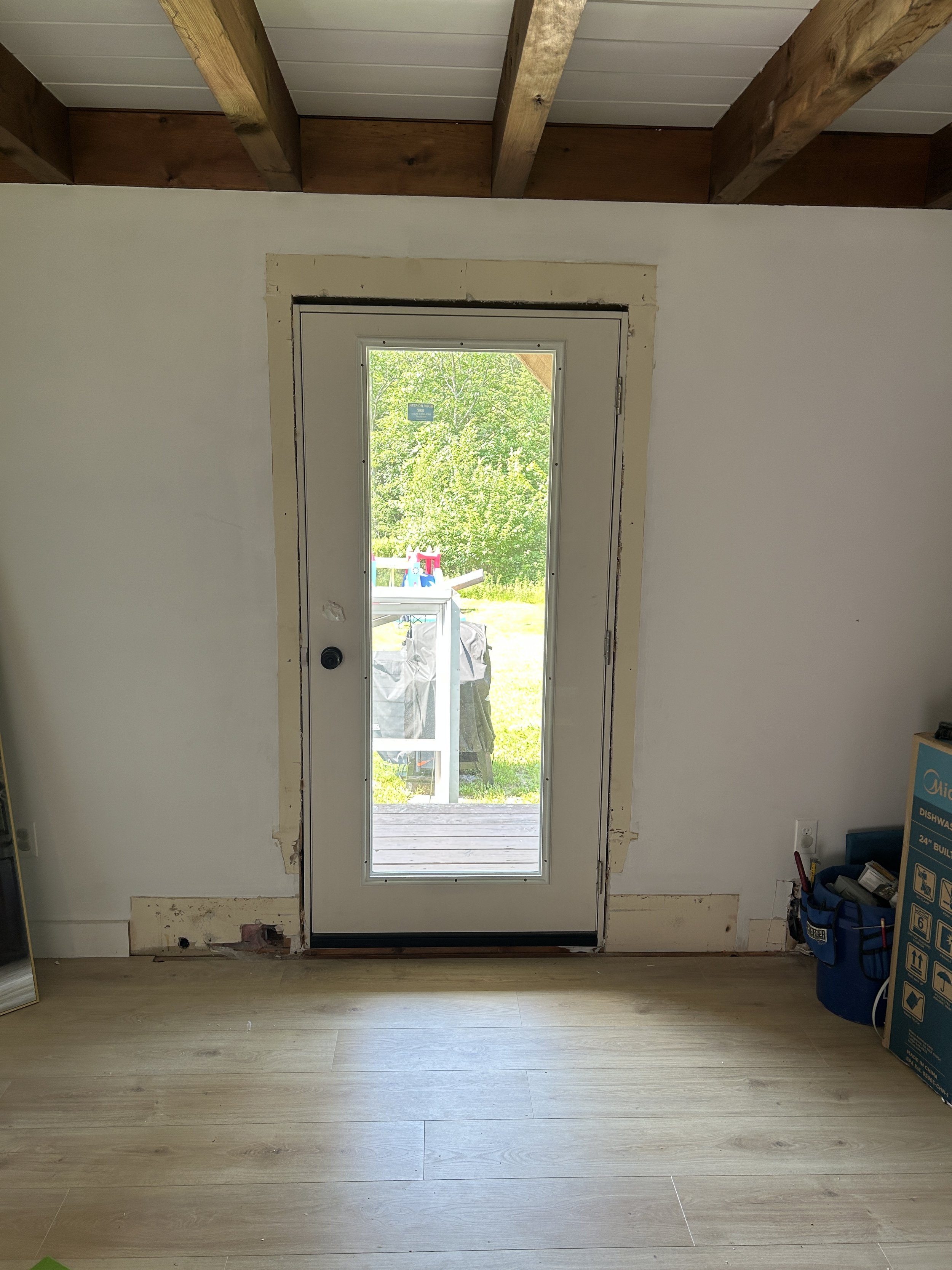 Interior view of a room with a door leading outside, unfinished wall framing around the door, and a wooden ceiling with exposed beams. Outside view shows trees and a playset in a yard.