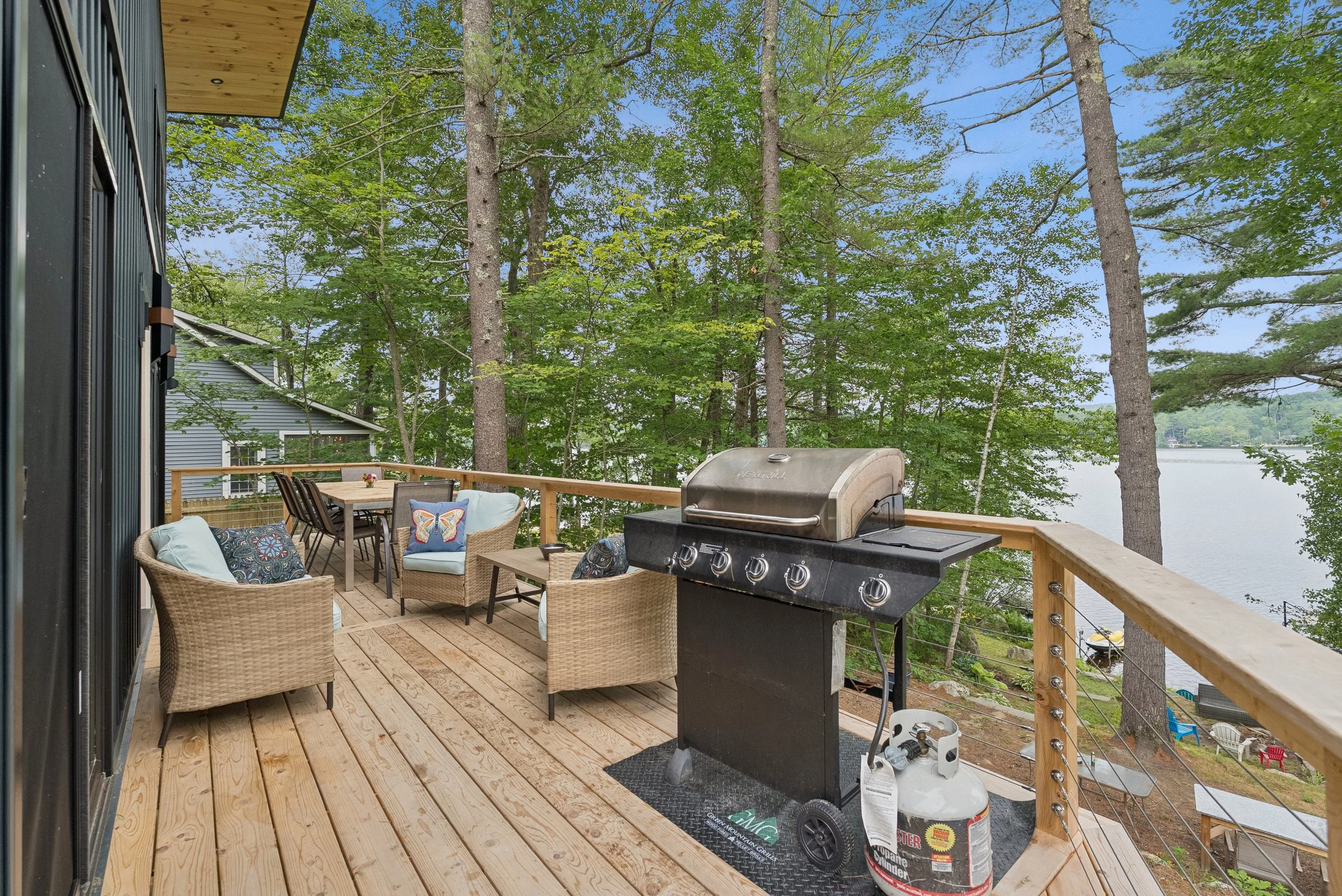 A wooden deck with outdoor furniture including beige wicker chairs with cushions and a dining table with chairs. There is a barbecue grill on the deck. The deck has a railing made of wooden posts and cables. Beyond the deck, there are tall green tree