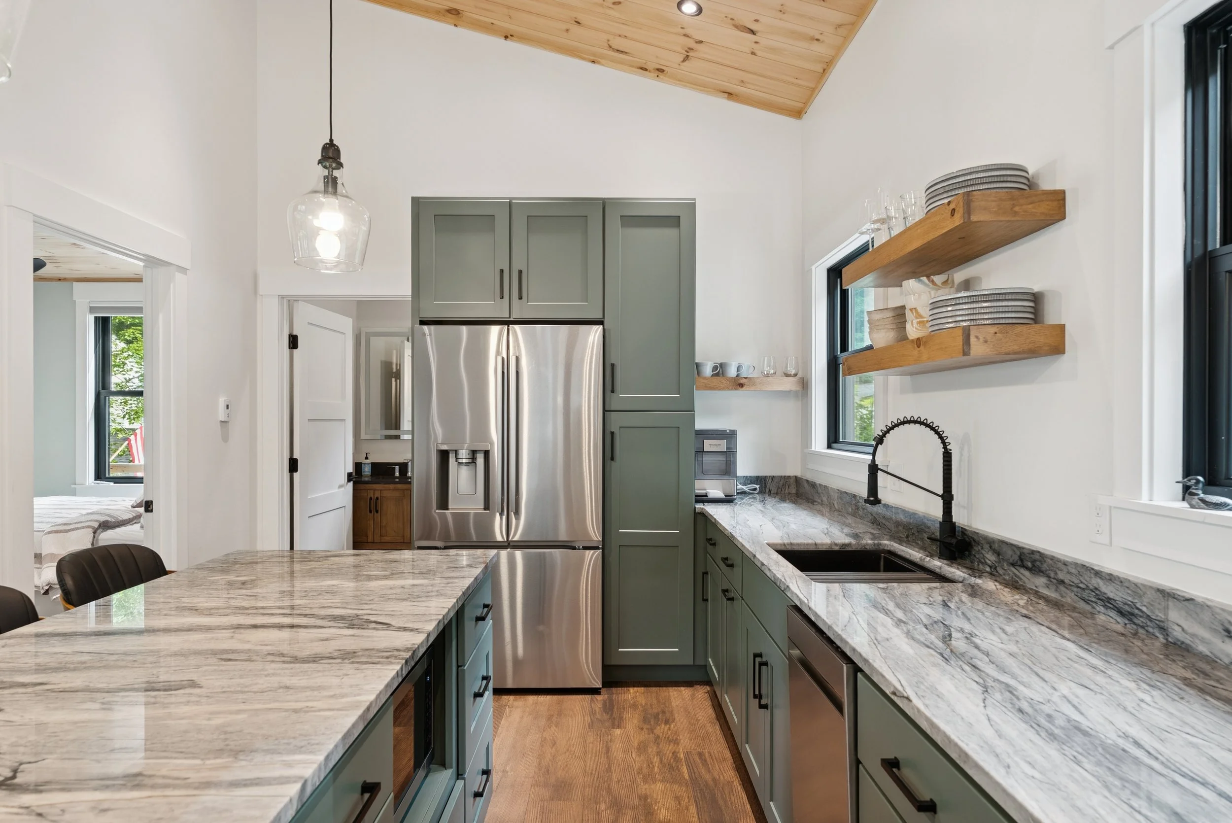 Modern kitchen with green cabinets, marble countertops, stainless steel fridge, black sink with gooseneck faucet, wooden floating shelves with dishes, and a sloped wooden ceiling with recessed lighting.