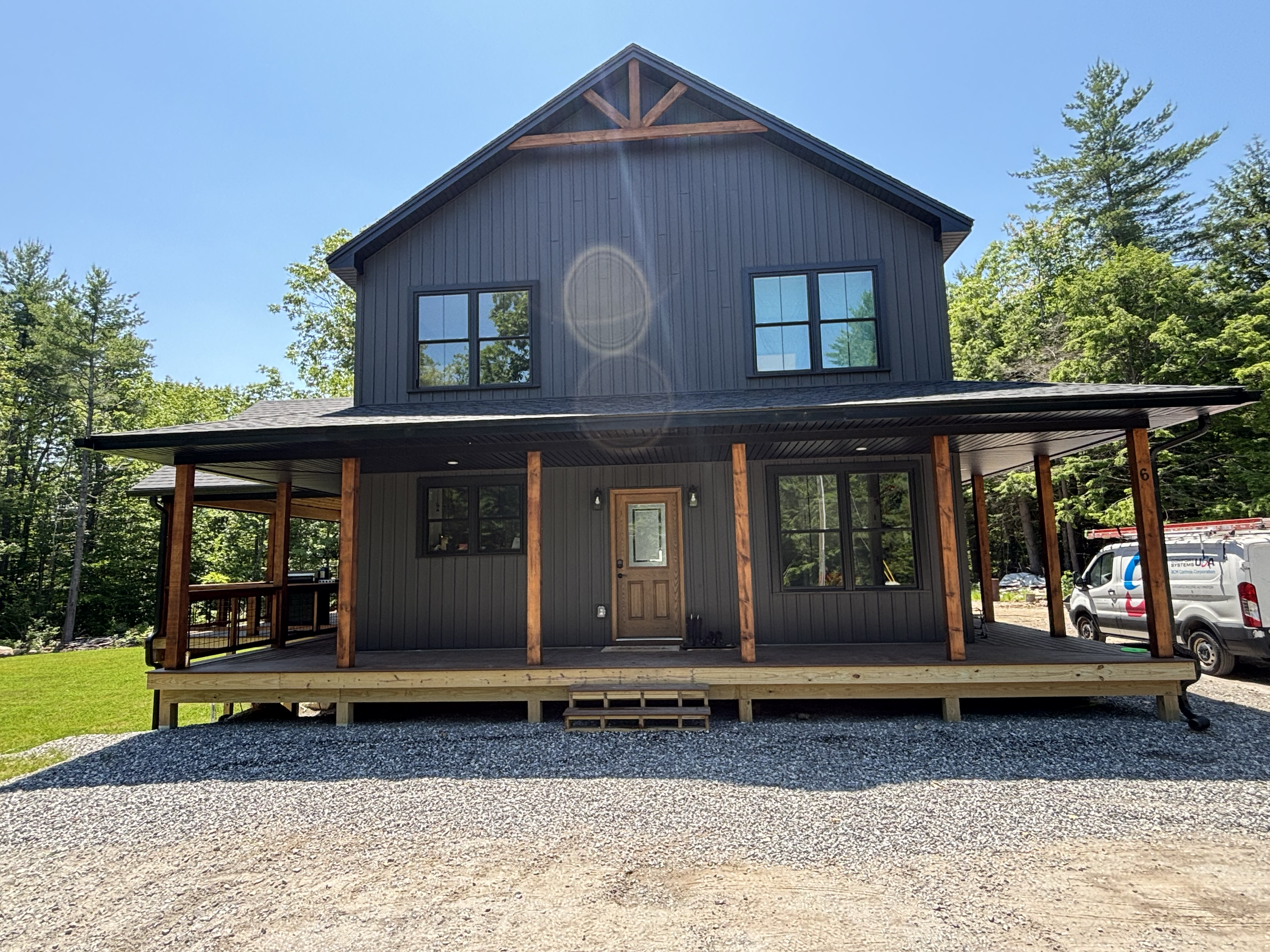 A two-story house under construction with a wooden porch, gray siding, and large windows, surrounded by green trees and a clear blue sky.