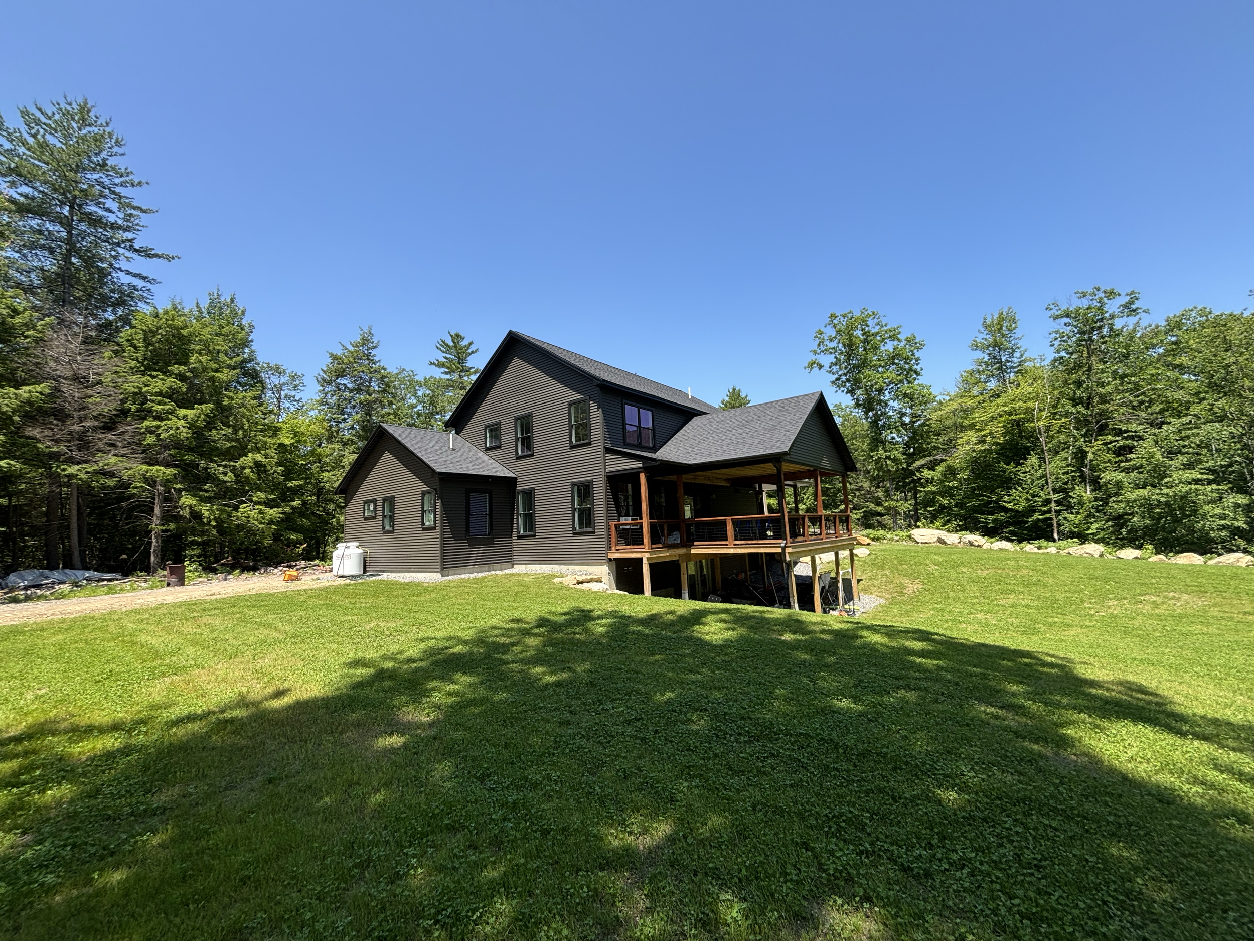 A two-story house with dark siding and a gray roof, surrounded by green trees and a well-maintained lawn under a clear blue sky.