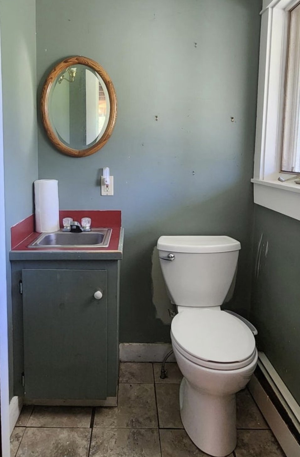 A small bathroom with green walls and a tile floor, containing a toilet and a small sink with a red countertop, a mirror with a wooden frame, and a window on the right side.
