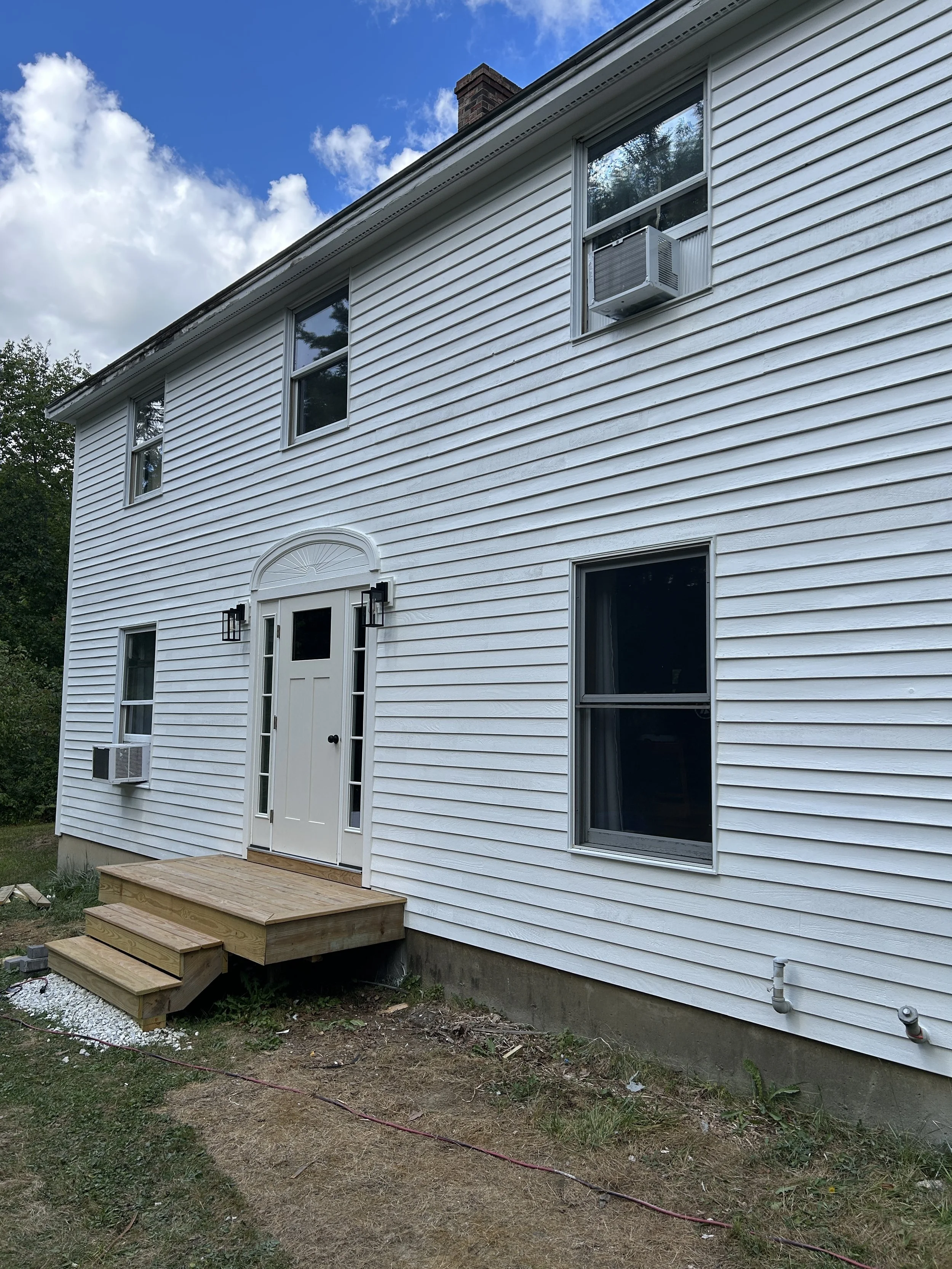 Back of a white house with vinyl siding, three windows, a door with glass panel, and a small set of wooden stairs leading to the door. There is a brick chimney, a window air conditioning unit, and a partially green yard with dirt, grass, and some con