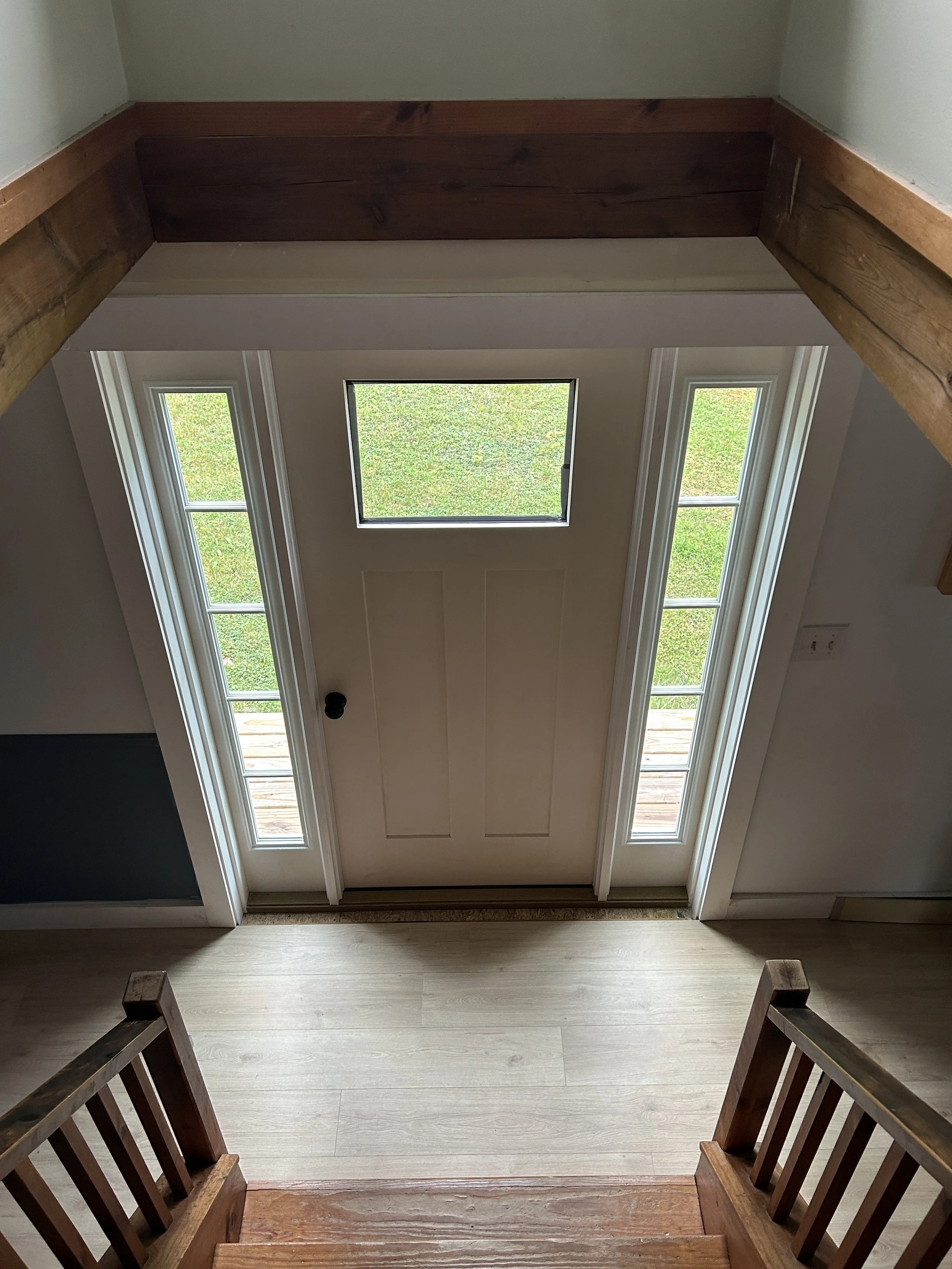 View from the top of a staircase looking down at a white entry door with a window above, with two sidelights and green grass outside. The staircase has wooden railings.