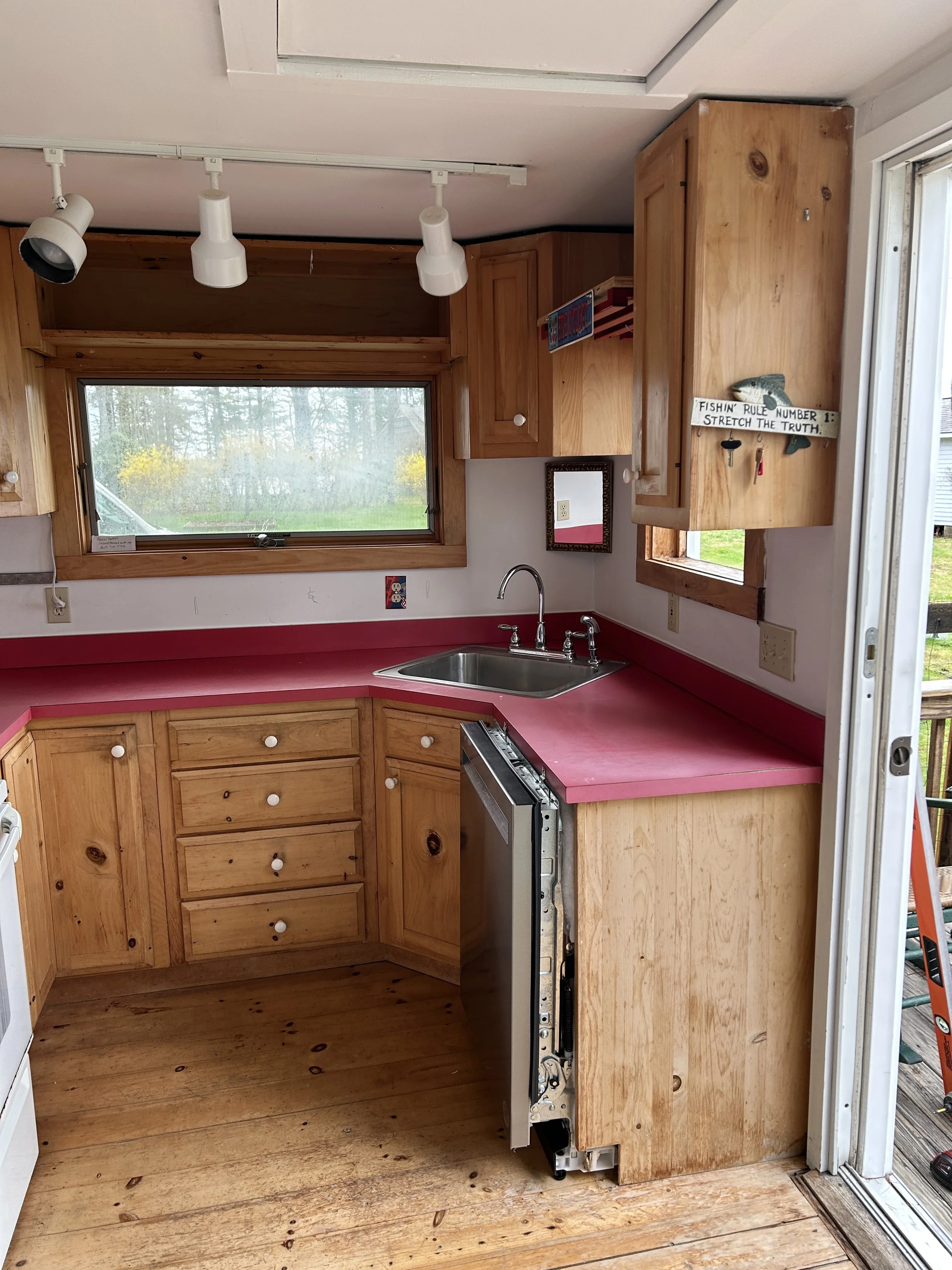 A partially built wooden kitchen with a window, a red countertop, a sink, and wooden cabinets, with a sign on the wall that reads, "Fishin' Rule Number 1: Stretch the Truth."