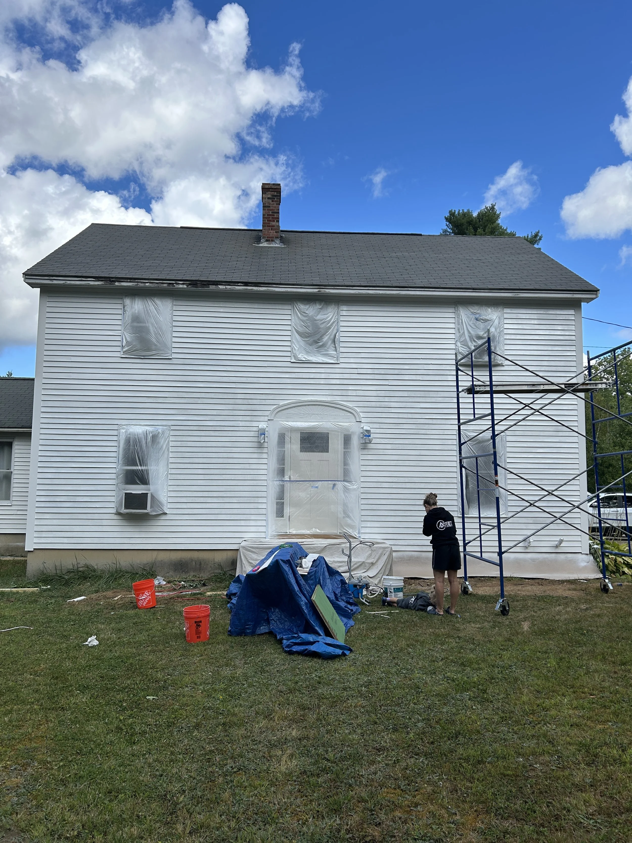 A two-story white house undergoing exterior renovation. The windows are covered with plastic sheeting, and scaffolding is set up on the right side. A person is standing near the scaffolding on the ground, and various tools and supplies are scattered 