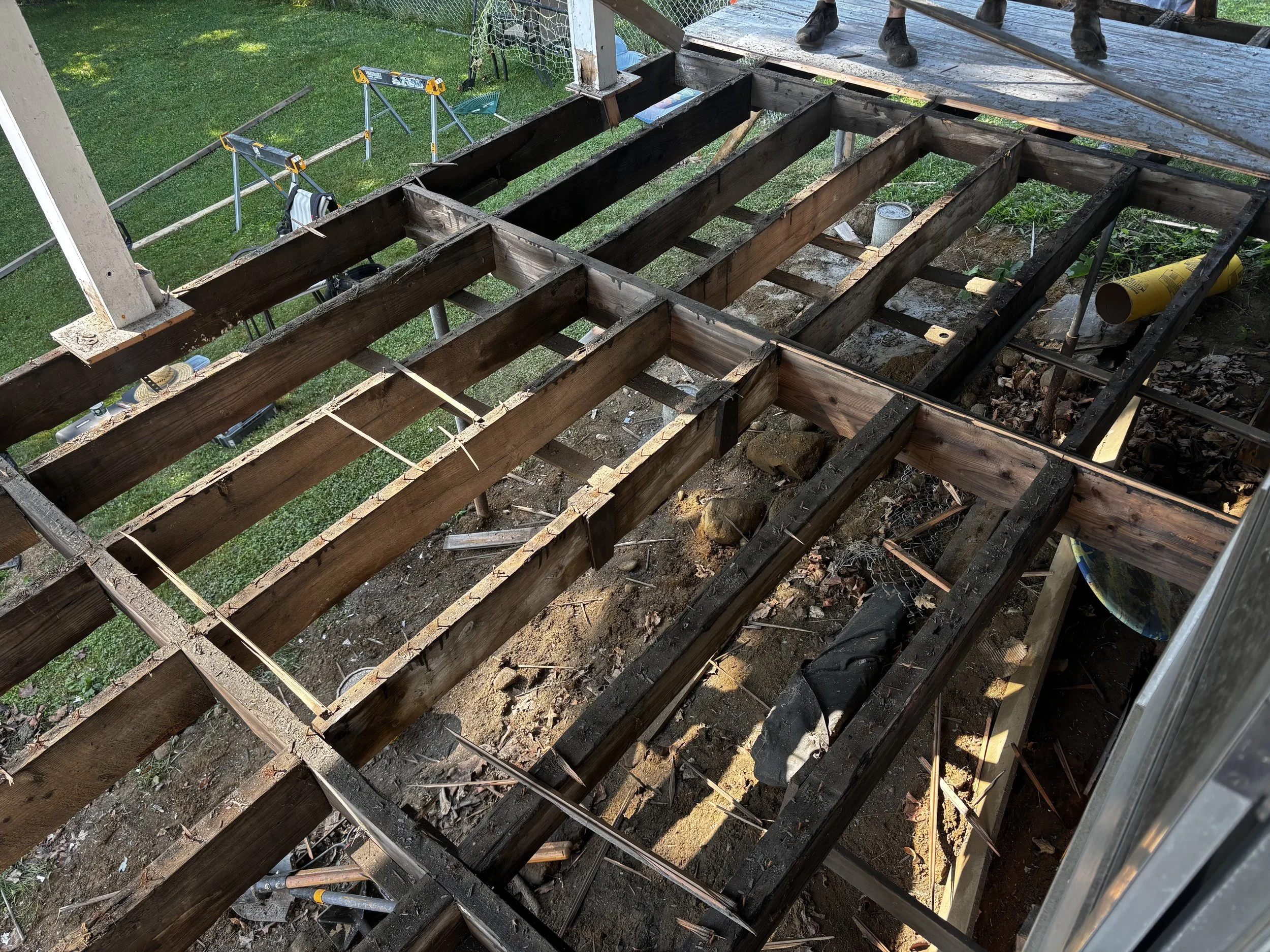 Construction site with exposed wooden framing for a deck renovation, with some tools and supplies visible, and a grassy backyard with playground equipment in the background.