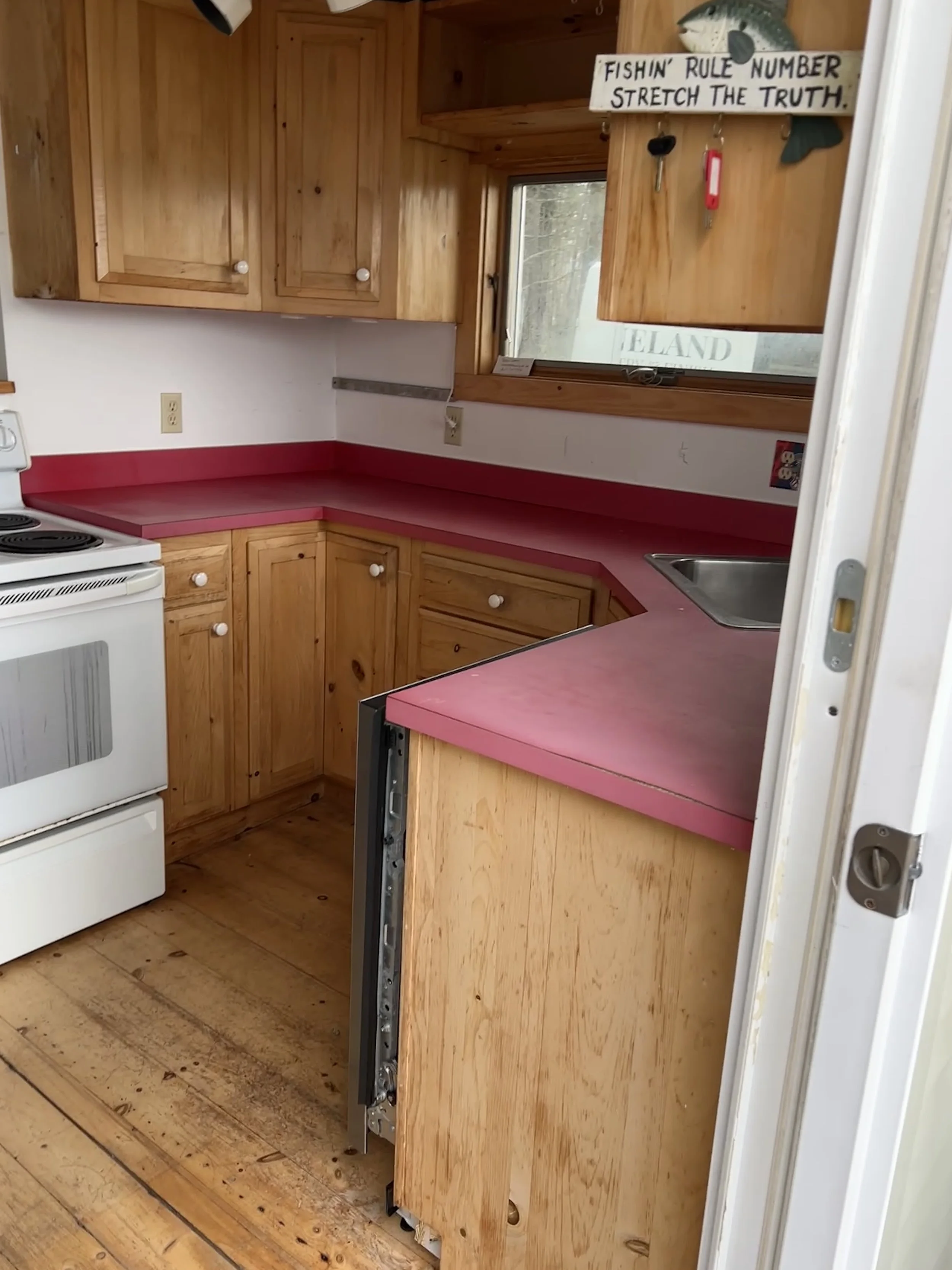 A small kitchen with wooden cabinets, a red countertop, and a window showing outdoors. There is a stove and an open space where a dishwasher might go, with faded wood flooring.