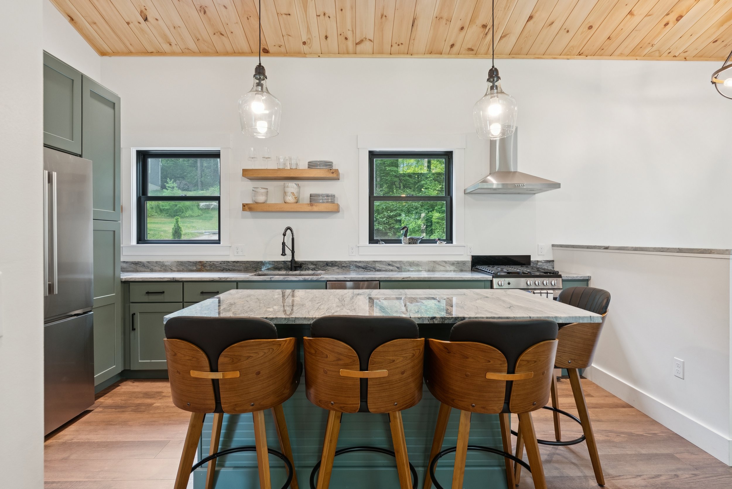 Modern kitchen with wooden ceiling, dual window views of greenery, green cabinetry, marble countertop, and six wooden chairs around a kitchen island.