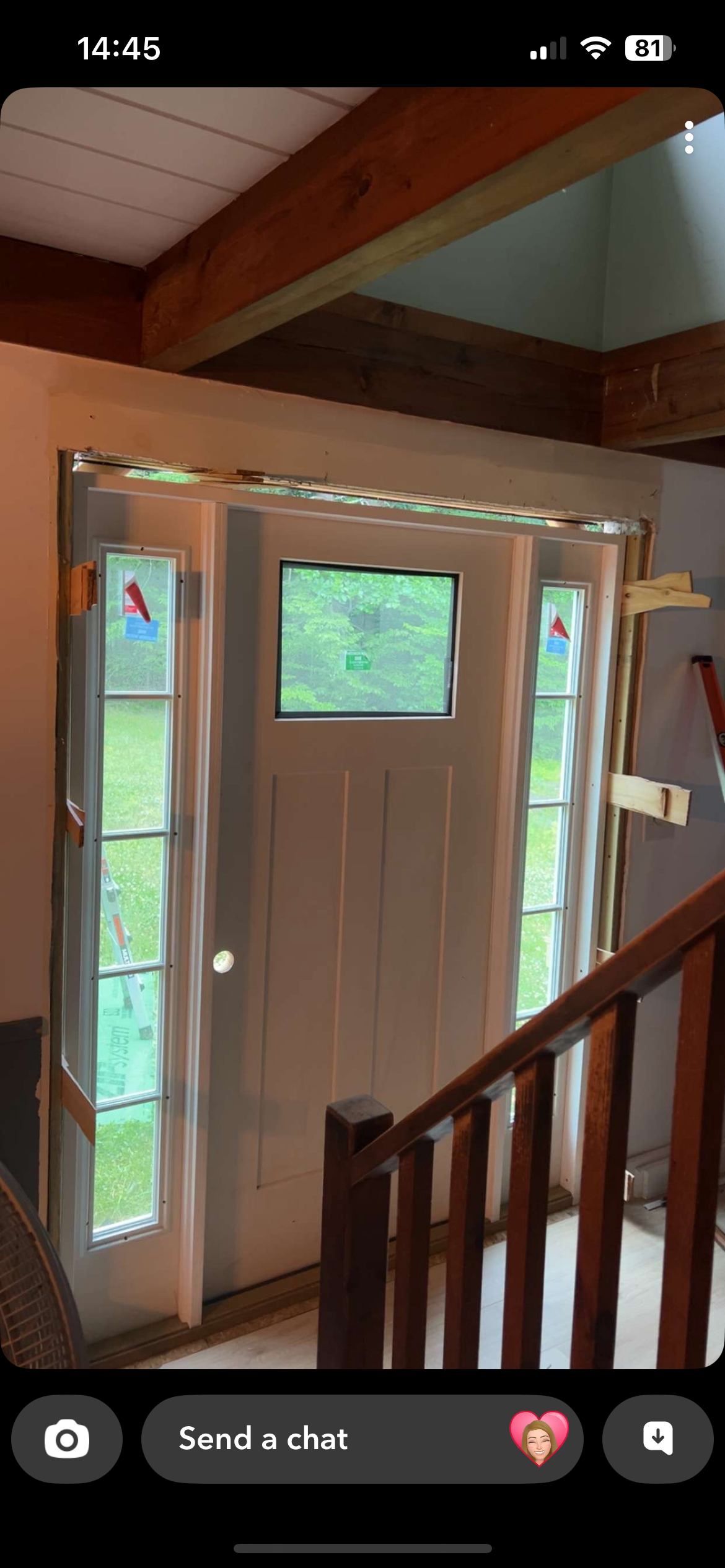View of a front door with sidelights and a small window above, under construction with exposed framing and a ladder outside visible through the sidelights. Wooden beams and stair railing are in the foreground.