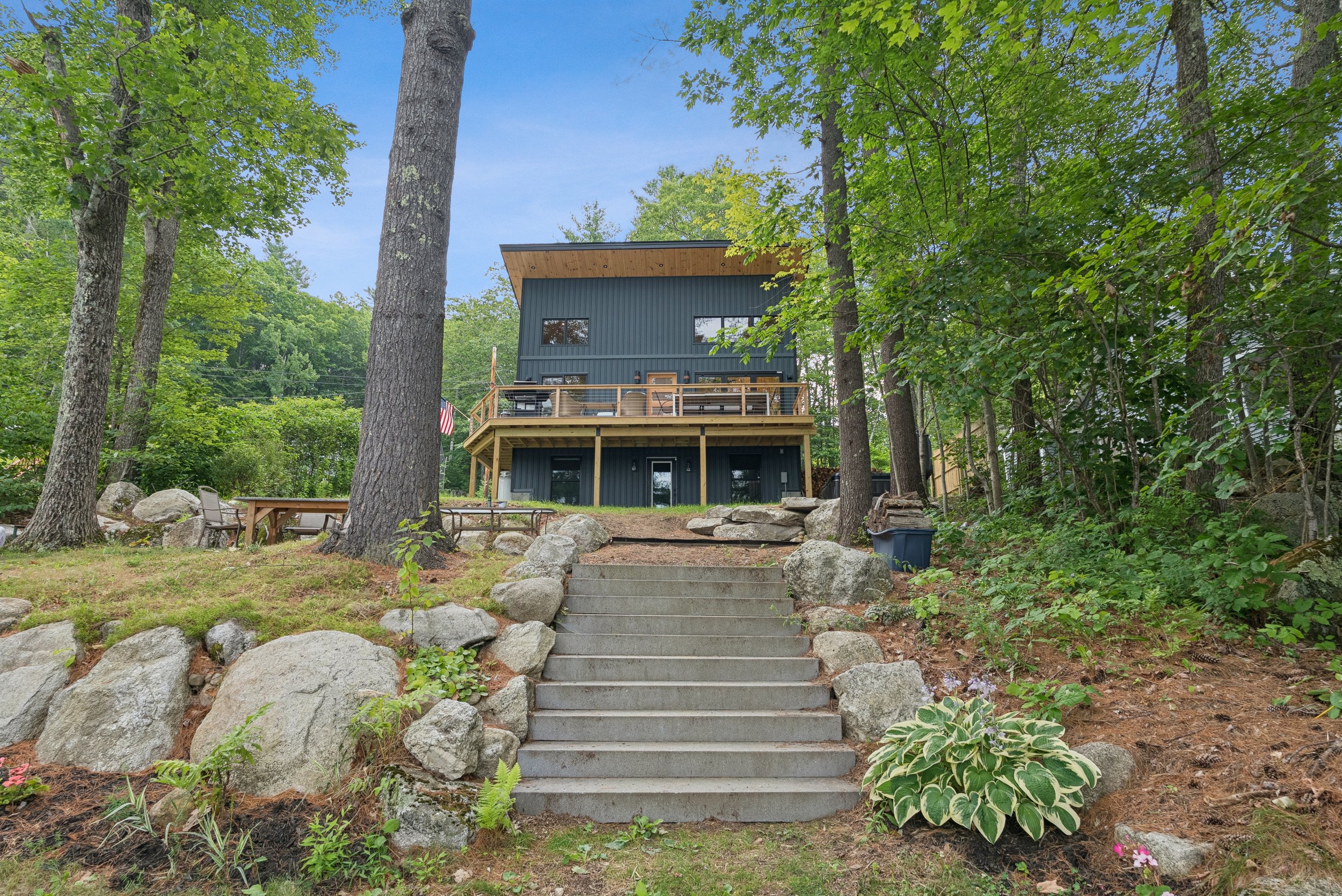 A two-story dark blue house with a wooden deck on the upper level, surrounded by tall green trees, with stone steps leading up to the deck and a garden area with rocks and plants in the foreground.
