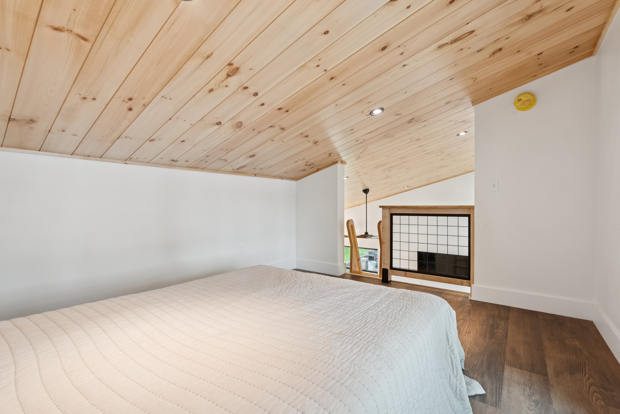 Interior of a bedroom with a wood-paneled ceiling, a white wall, a bed with a beige quilt, a fireplace, and a small window near the floor.